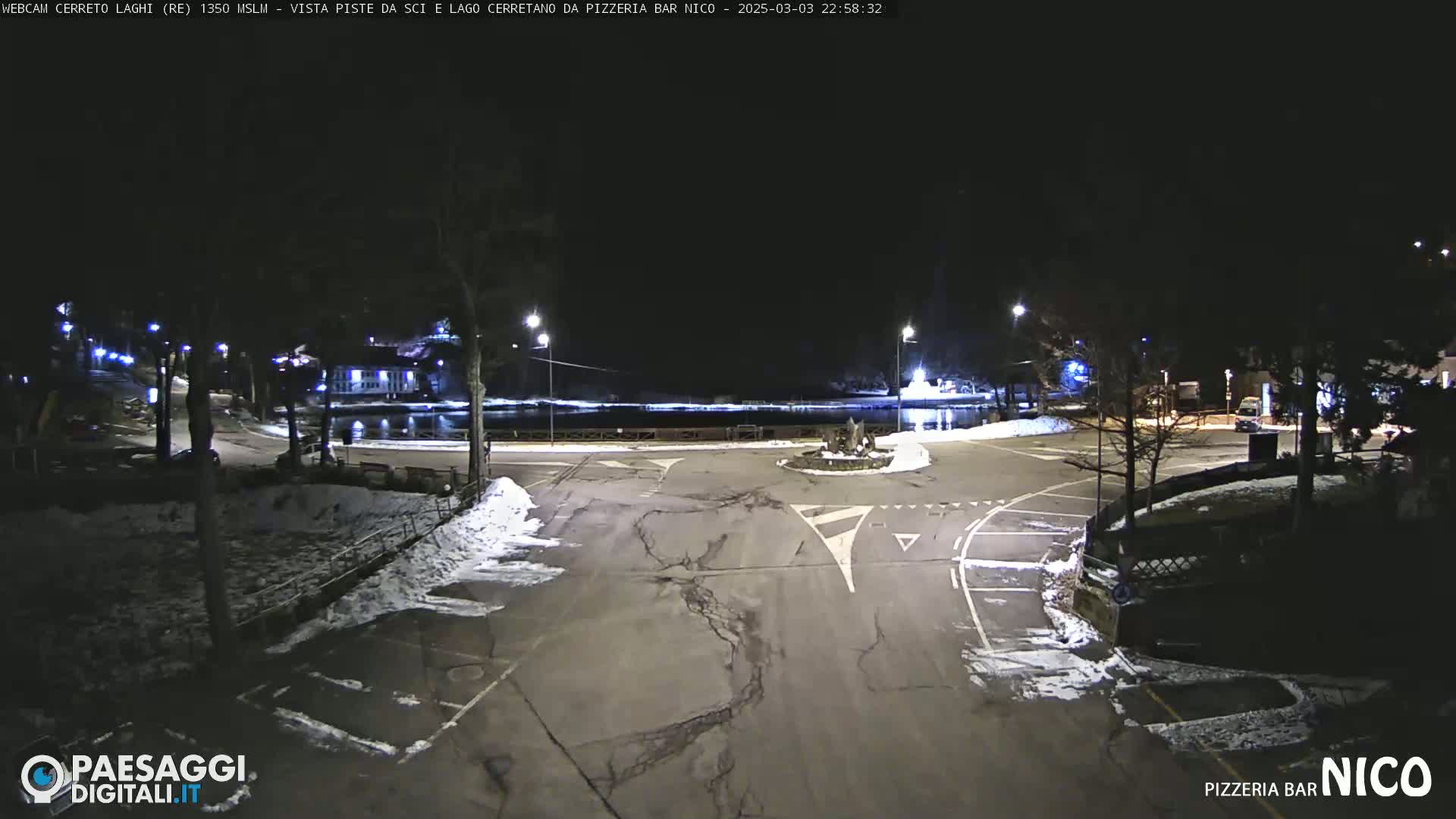 A nighttime view of a snow-dusted parking lot and roadway near a body of water, illuminated by streetlights.