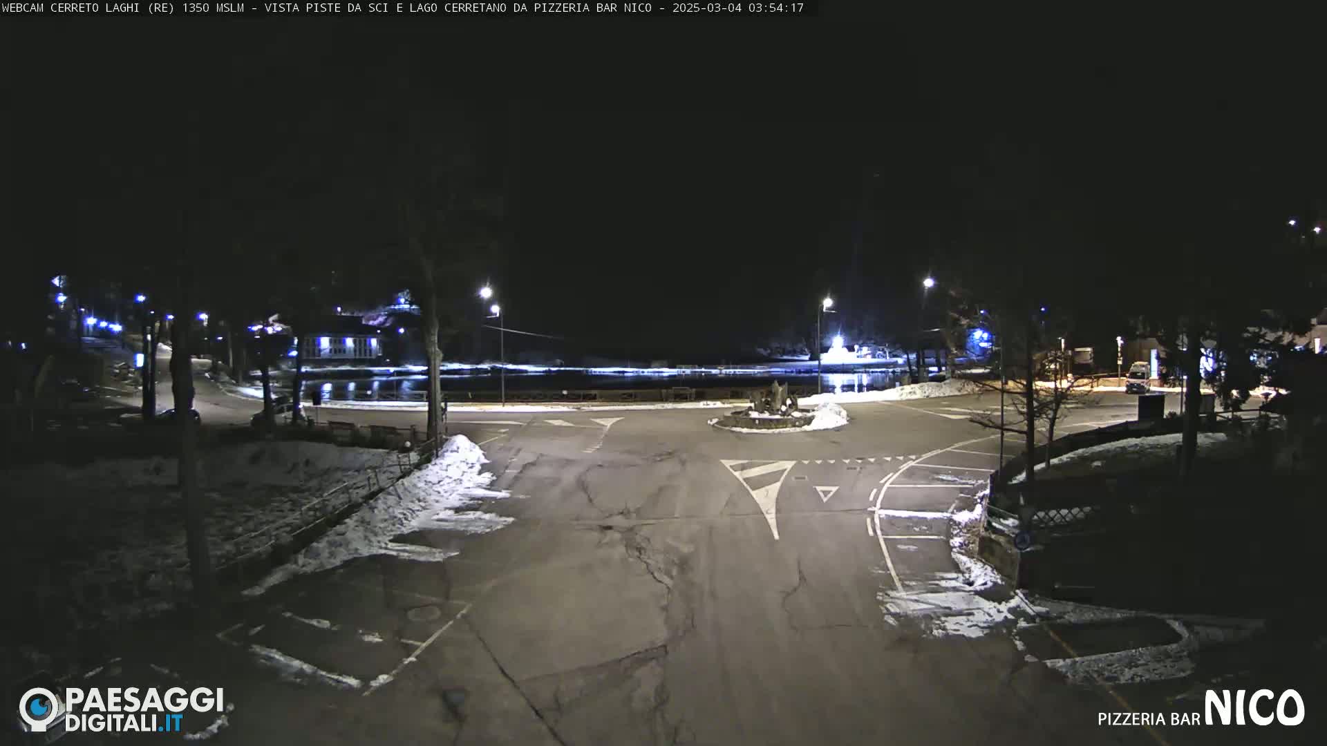 A nighttime view of a snow-dusted parking lot and roadway intersection near a body of water, illuminated by streetlights.