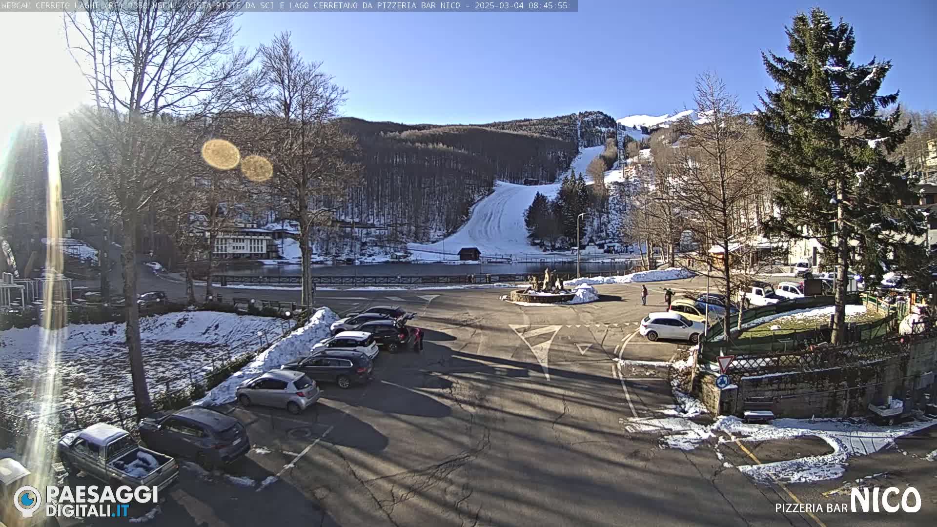 A sunny parking lot with several cars is situated in front of a snowy ski slope and wooded hillside.