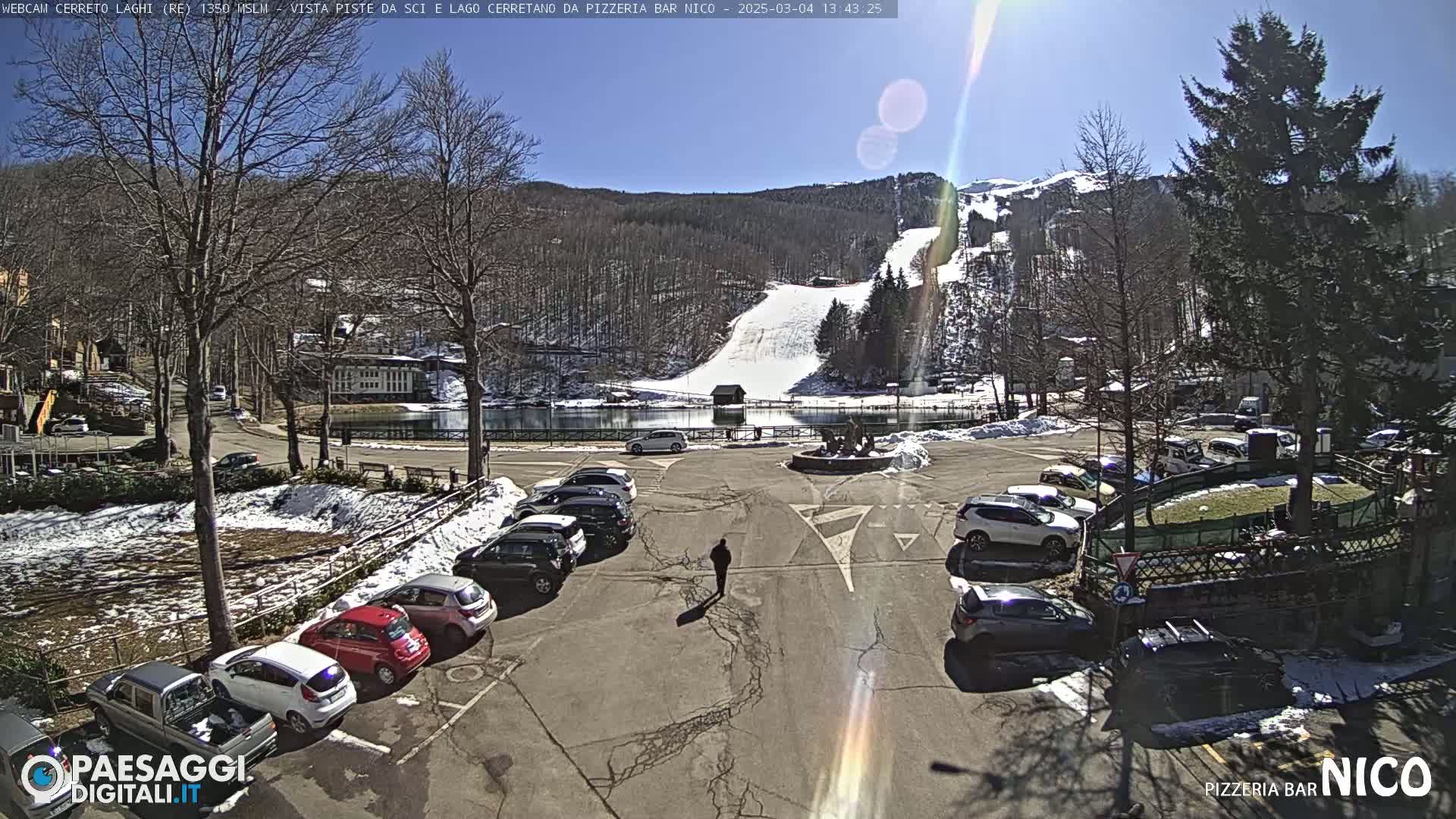 A sunny day in a mountain parking lot with some snow on the ground, showing a ski slope and a lake in the background.