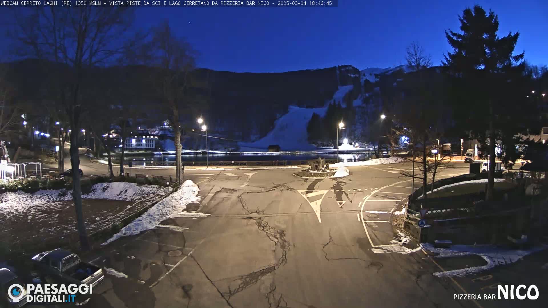 A nighttime, snow-dusted parking lot and roadway overlook a dark lake and snow-covered ski slopes under a twilight sky.