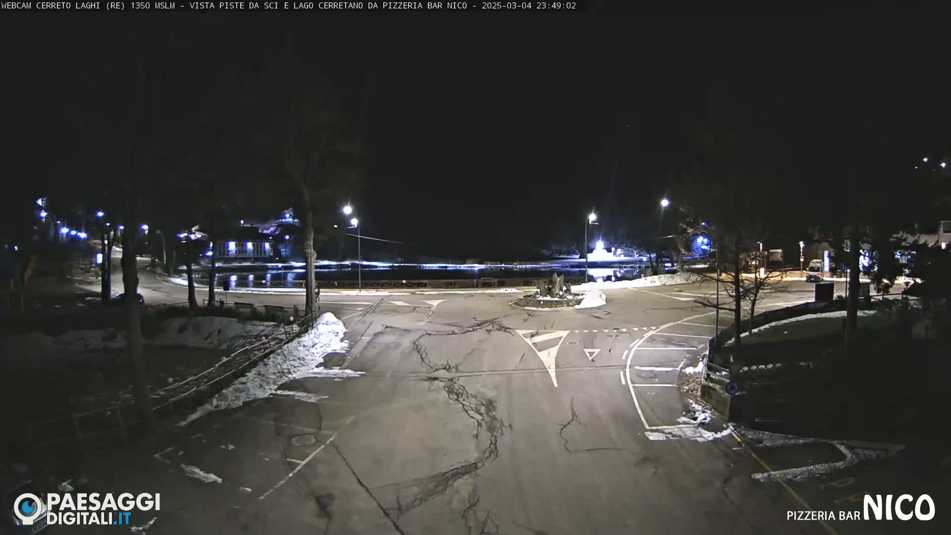 A nighttime view of a snow-dusted parking lot and roadway intersection, illuminated by streetlights, near a body of water.