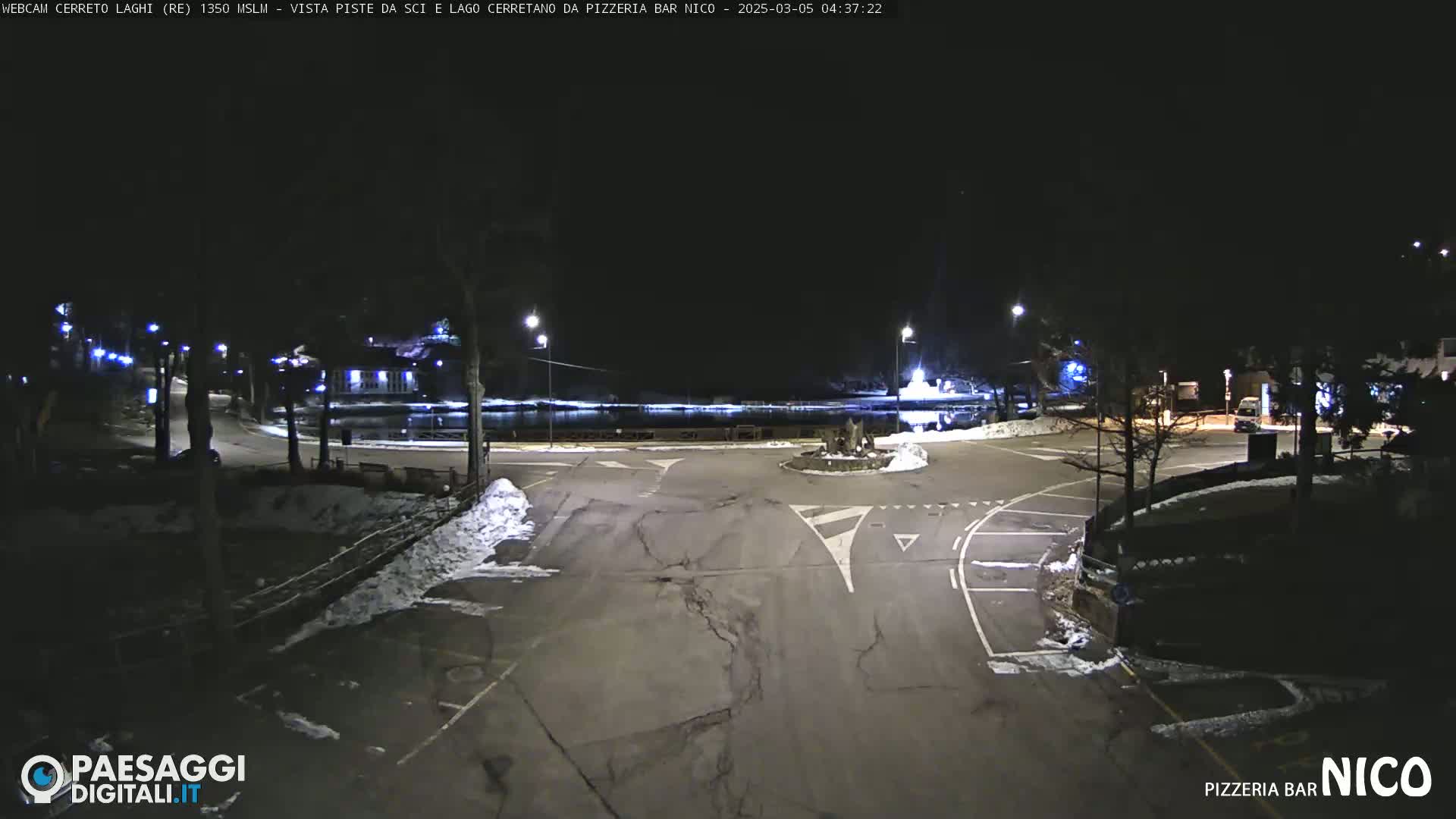 A nighttime view of a mostly empty parking lot and roadway intersection, with some snow on the ground and illuminated by streetlights.