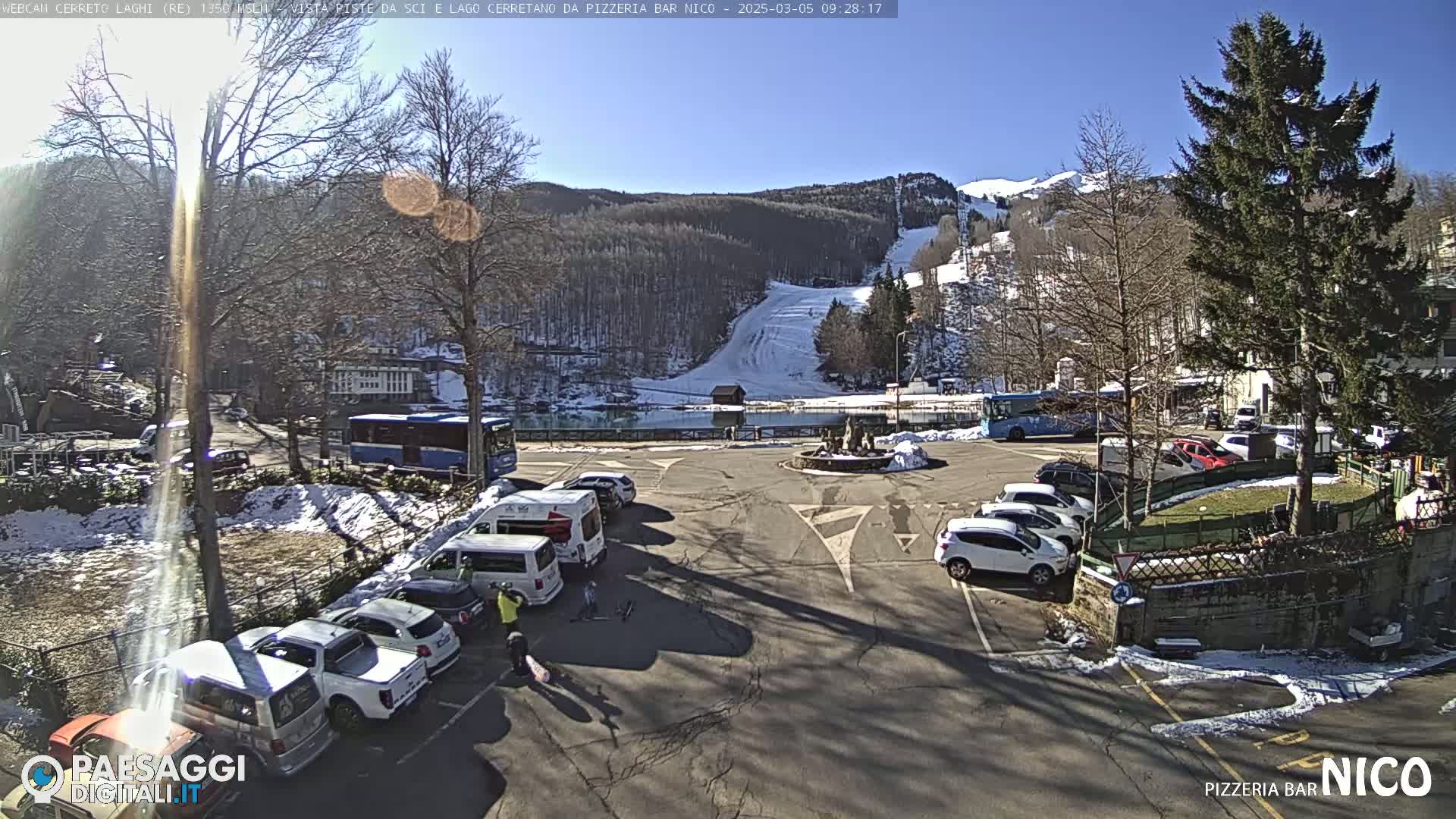 A sunny day in a mountain village with a parking lot full of cars and a ski slope visible in the background, some patches of snow are present.