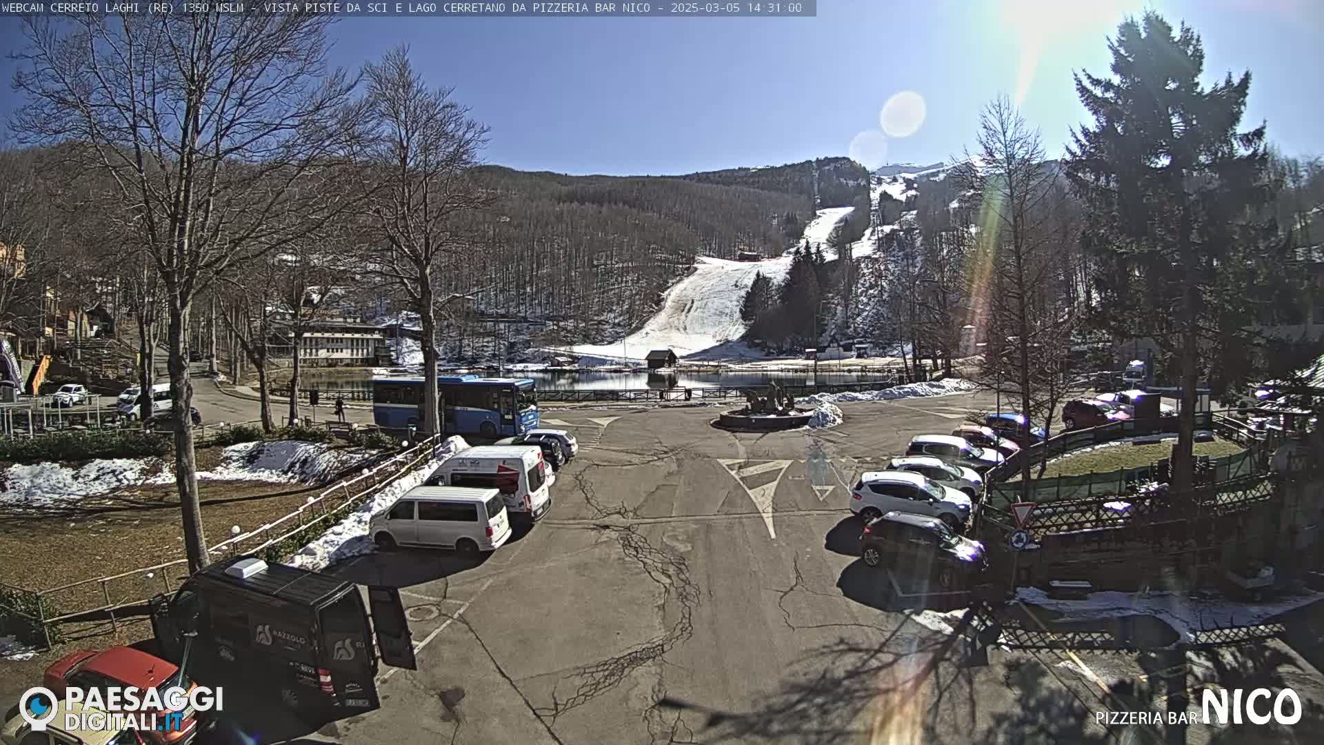 A sunny parking lot with several cars and buses, overlooking a snow-covered ski slope and a small lake, nestled among bare trees.