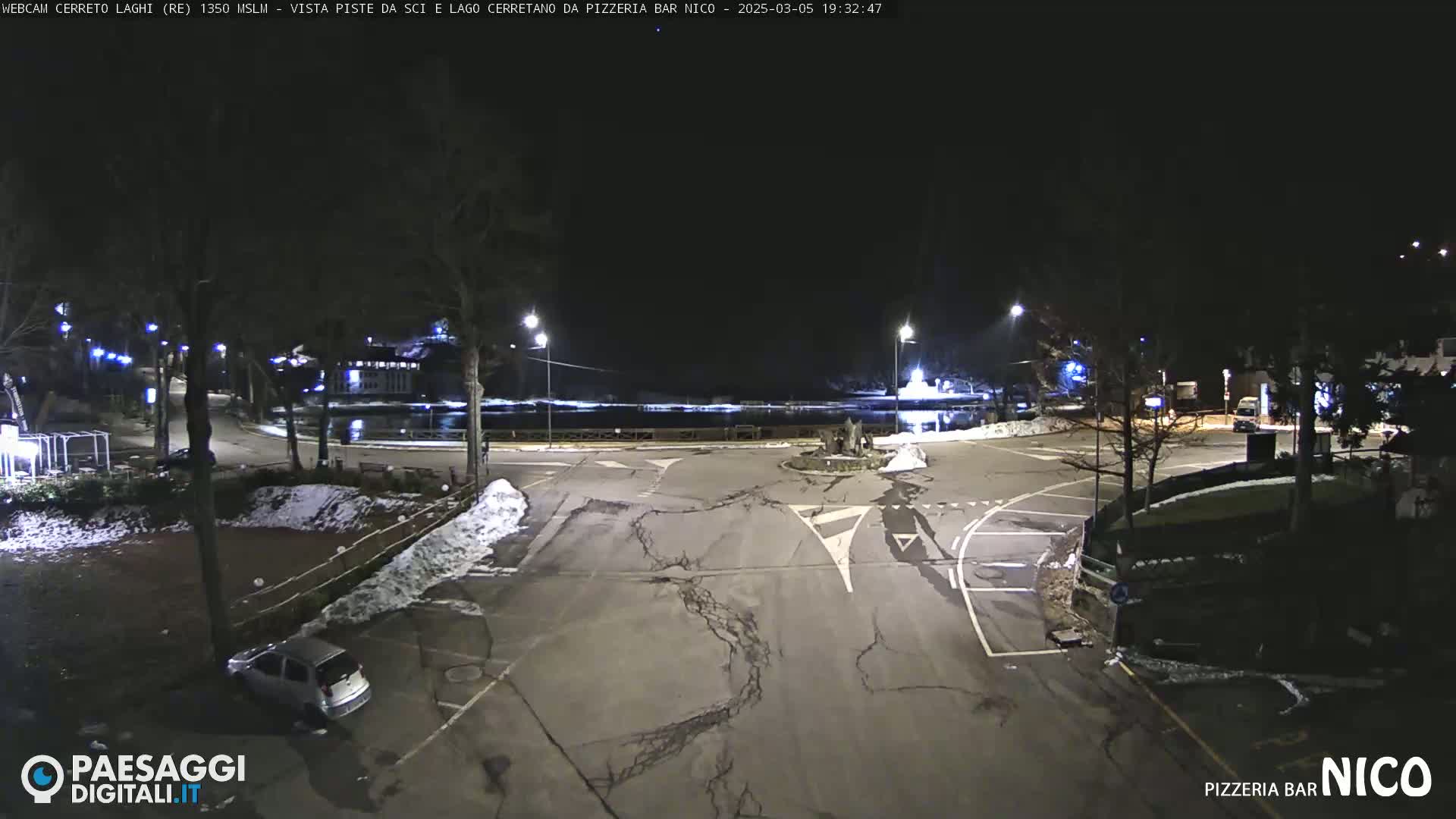 A nighttime view of a mostly empty parking lot and roadway near a body of water, with some snow on the ground and sparsely lit trees.