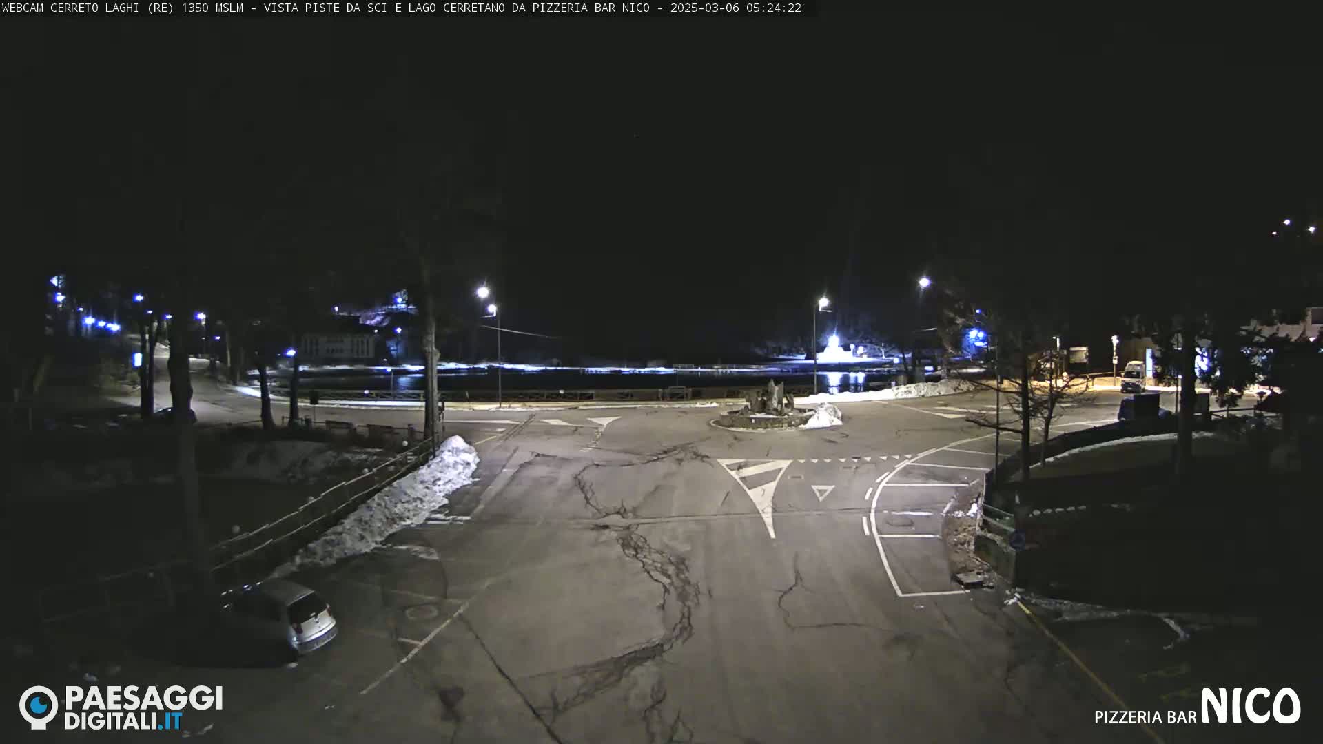 A nighttime view of a parking lot and roadway near a body of water, with some snow on the ground and sparsely lit trees in the background.