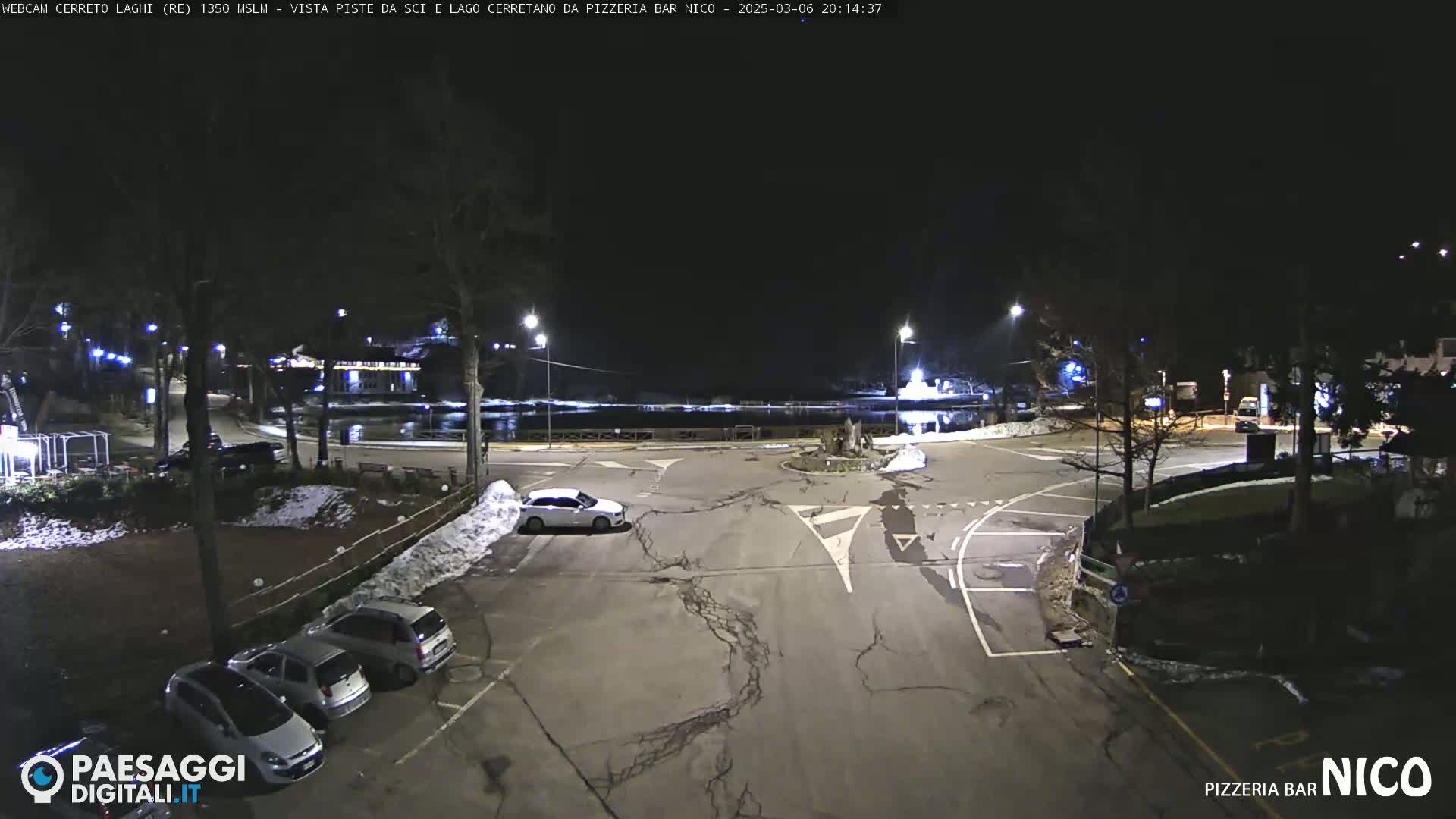 A nighttime view of a parking lot and roadway near a body of water, with several cars parked and some snow on the ground.
