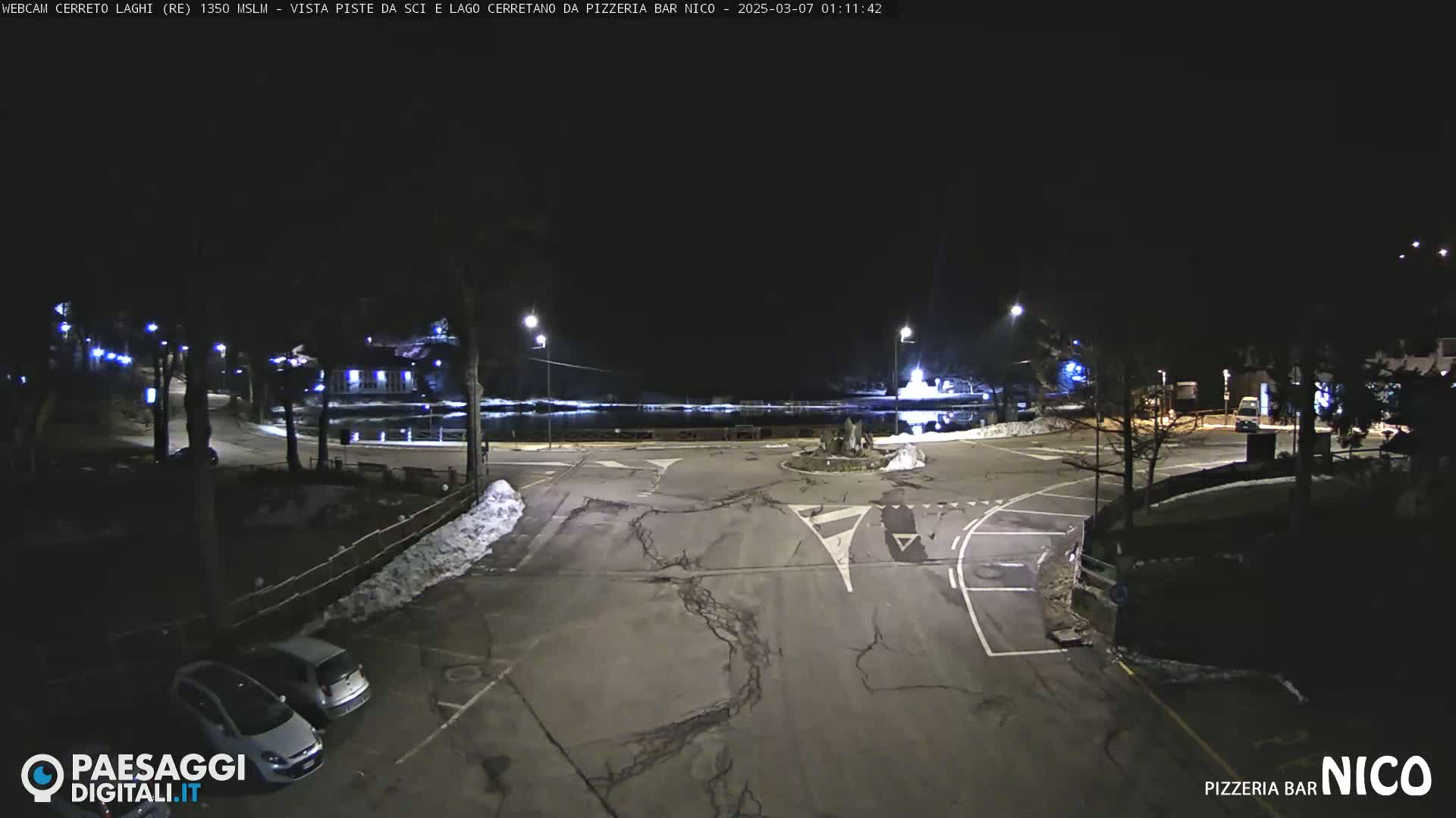 A nighttime view of a parking lot and road intersection with some cars parked, partially snow-covered ground, and streetlights illuminating the scene.