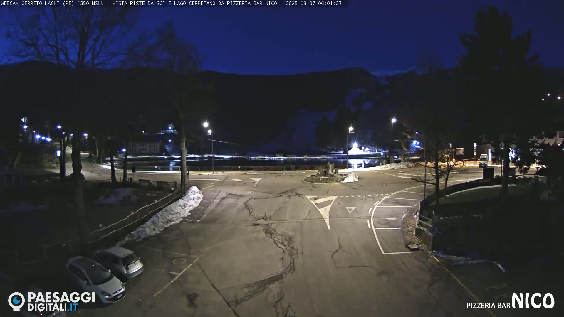 A nighttime view of a mostly empty parking lot and roadway intersection near a lake, with some snow on the ground and dark mountains in the background under a clear, dark sky.