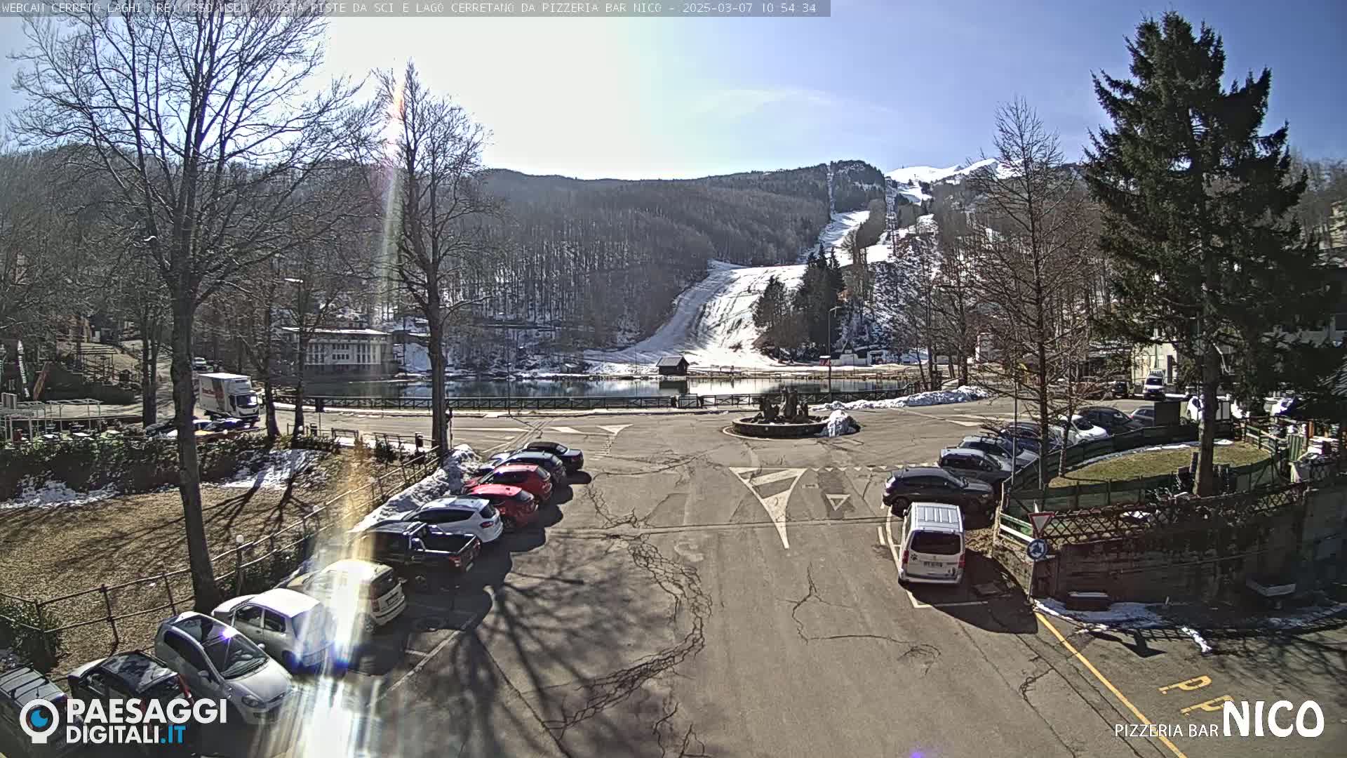 A sunny day shows a parking lot with cars, a small lake, and a snow-covered ski slope in the background.