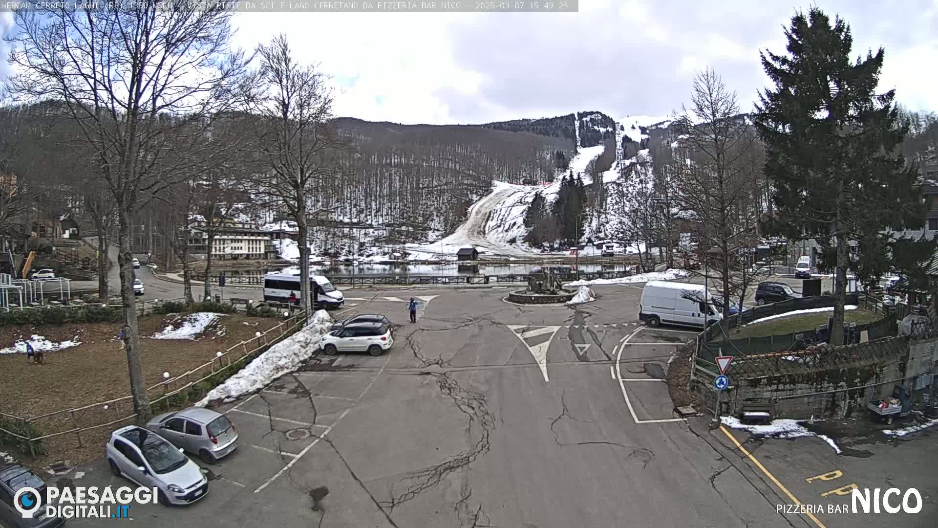 A parking lot with several cars, vans, and a small building is situated in front of a snow-covered ski slope and a lake, under a partly cloudy sky.