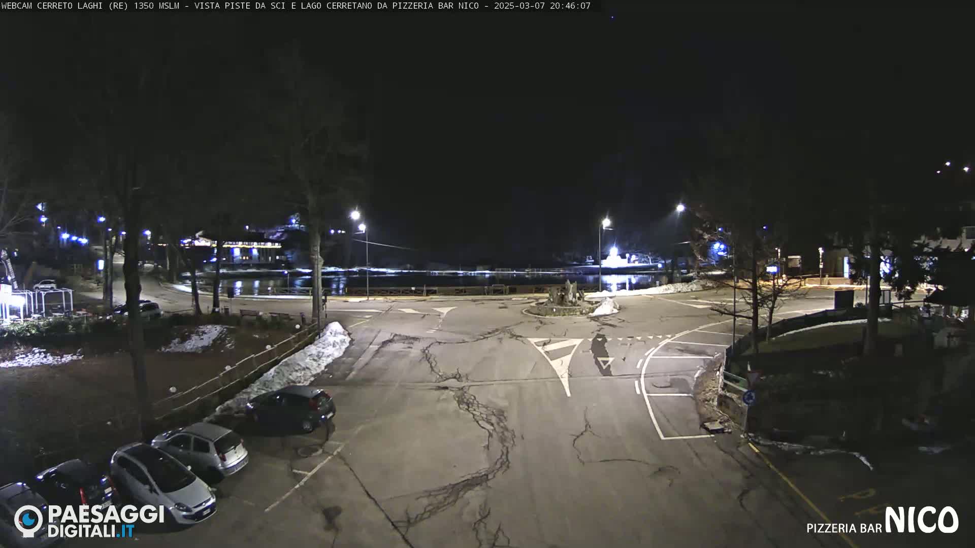 A nighttime view of a parking lot and roadway near a body of water, with some snow on the ground and cars parked.