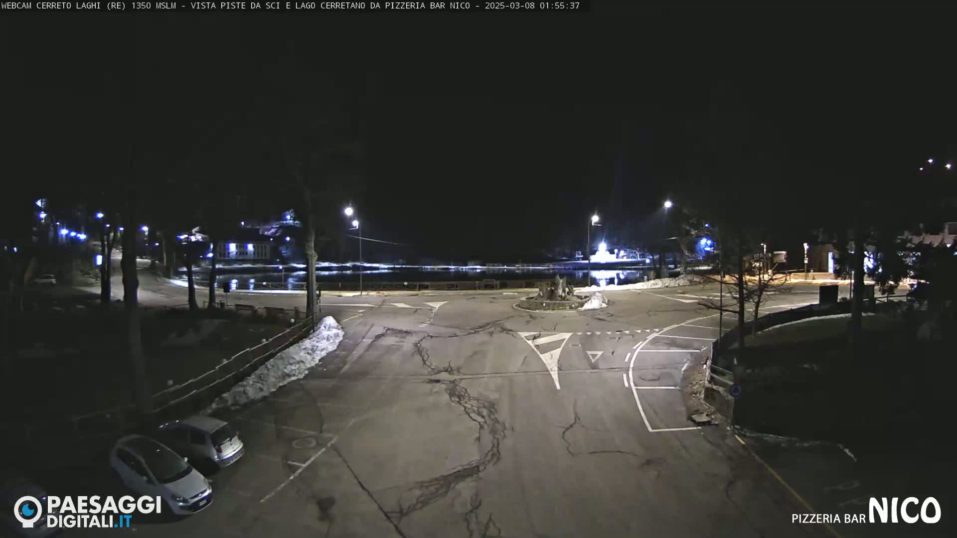 A nighttime view of a mostly empty parking lot and roadway intersection near a body of water, with some snow on the ground and a few cars parked.