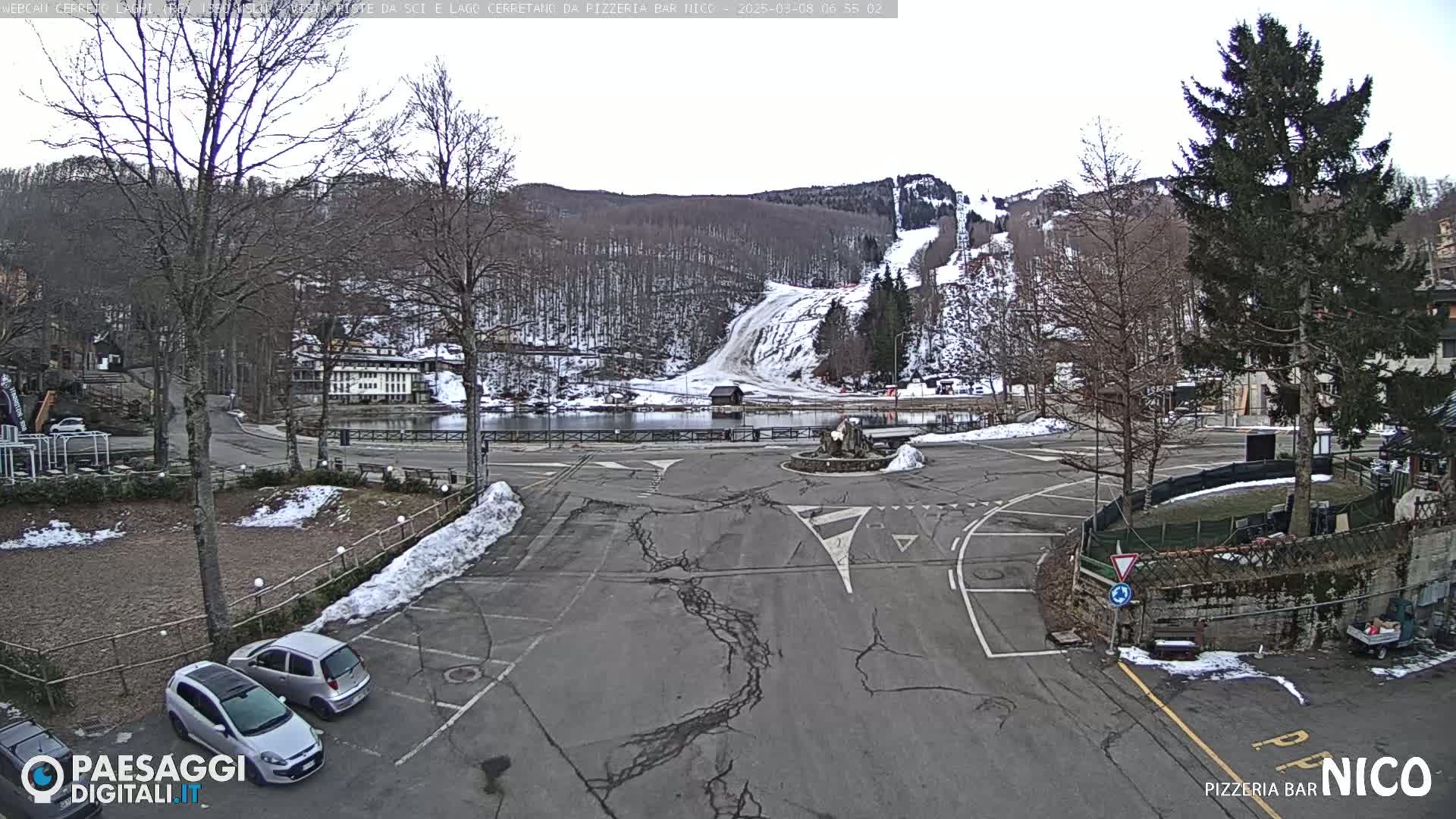 A mostly empty parking lot with a few cars, surrounded by trees and a small body of water, with a snow-covered ski slope visible in the background under an overcast sky.