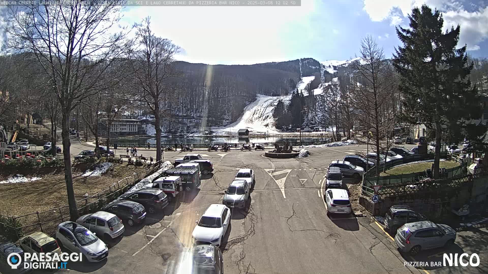 A sunny day reveals a parking lot full of cars overlooking a lake and ski slope nestled in a mountainside.