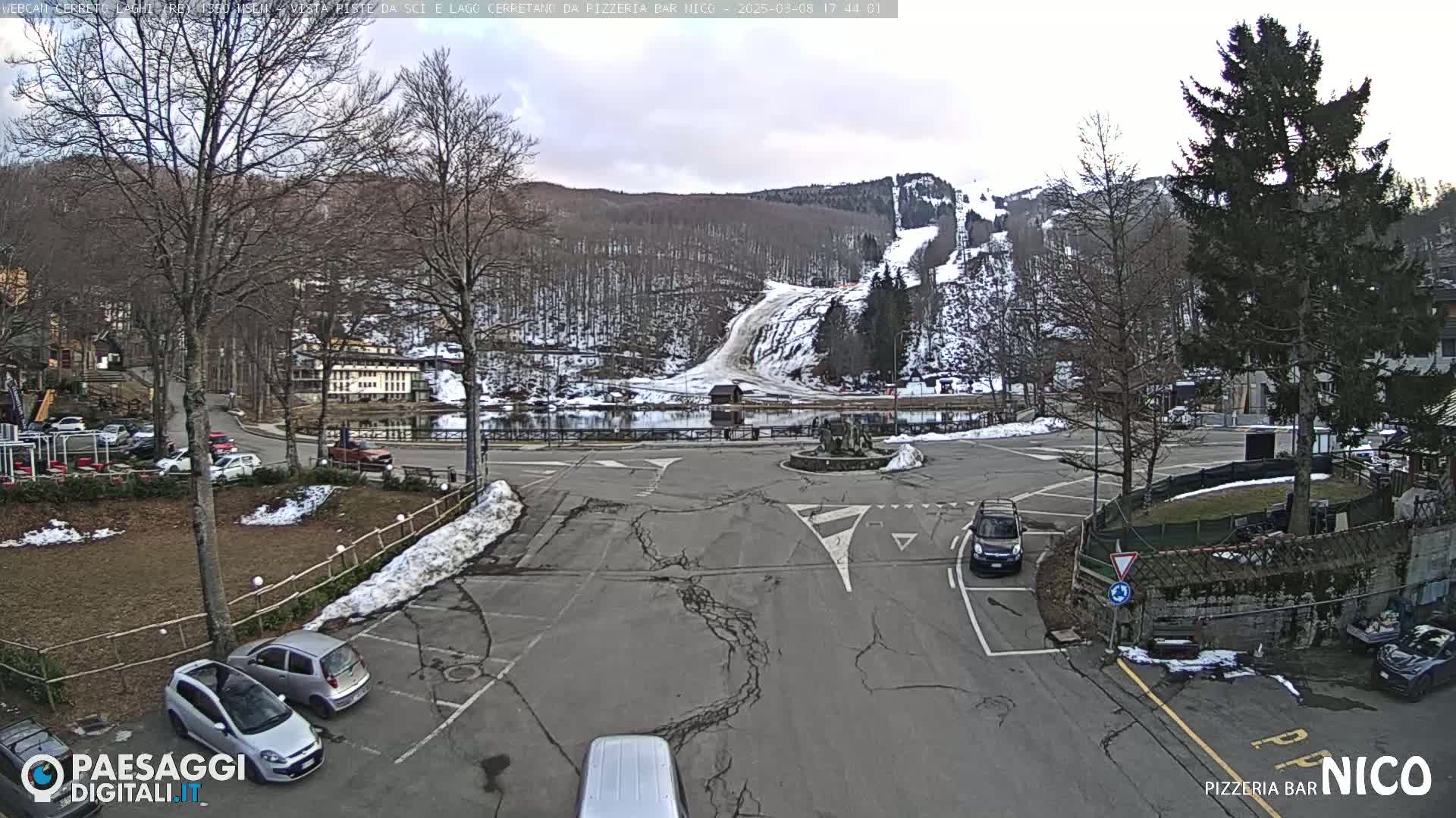 A parking lot with several cars and a view of a snow-covered ski slope and lake in the background under a cloudy sky.
