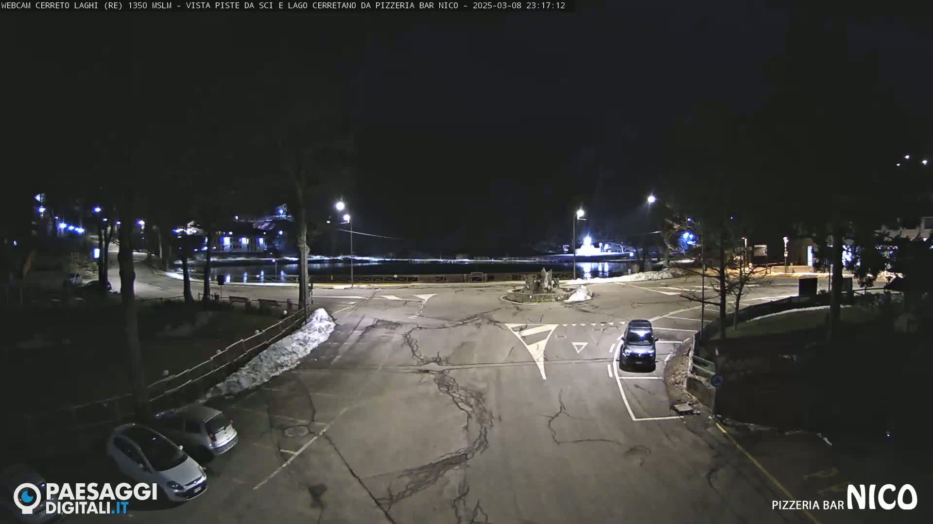 A nighttime, snow-dusted parking lot and roadway scene near a dark body of water, illuminated by streetlights.