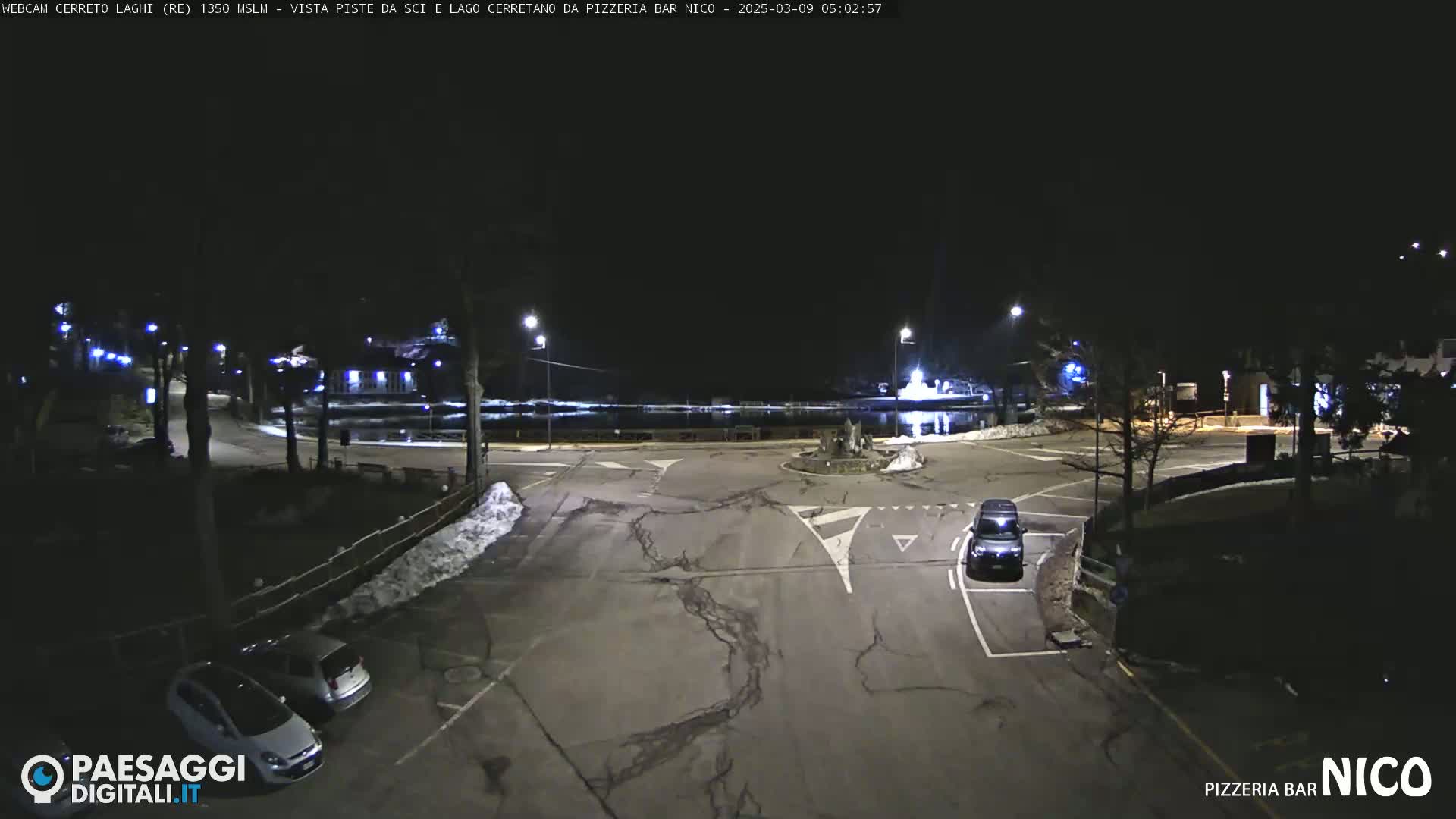 A nighttime, snow-dusted parking lot and roadway intersection are illuminated by streetlights, with a few cars visible.