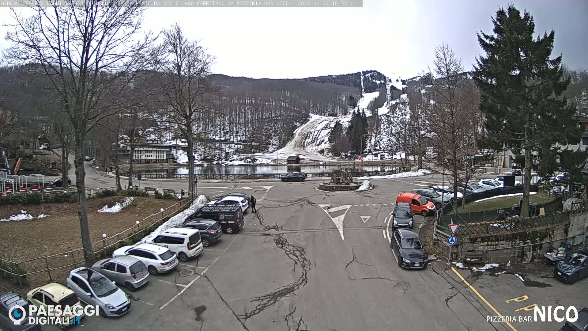 A parking lot with several cars is situated near a small lake and a snow-covered ski slope under an overcast sky.