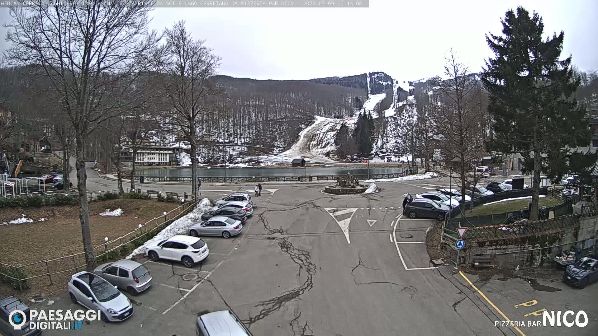 A parking lot with several cars is situated near a lake and ski slope under an overcast sky.