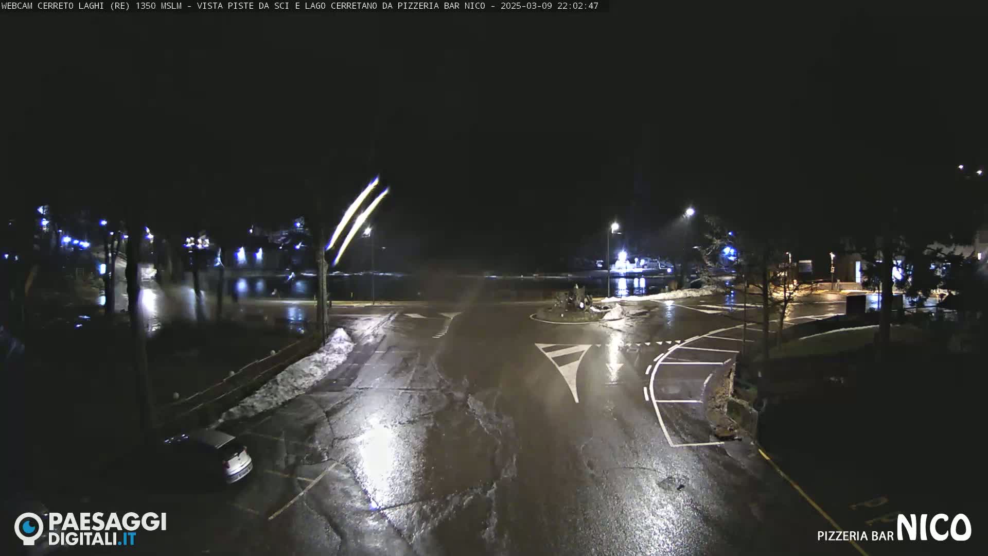 A nighttime view of a wet, empty parking lot and roadway near a body of water, with some snow visible on the ground and faint streetlights illuminating the scene.