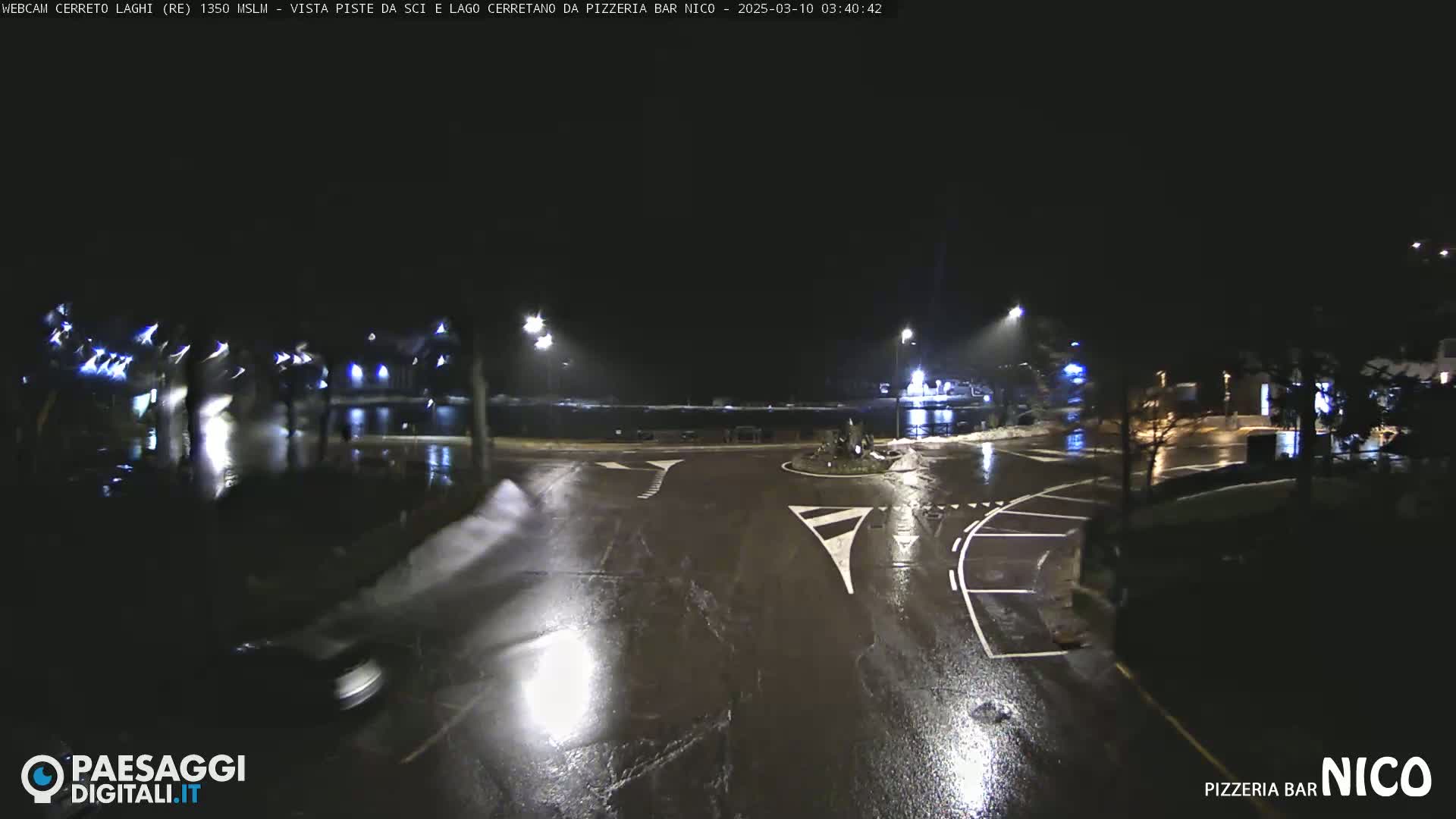 A wet, nighttime view of a roundabout and parking lot, illuminated by streetlights.