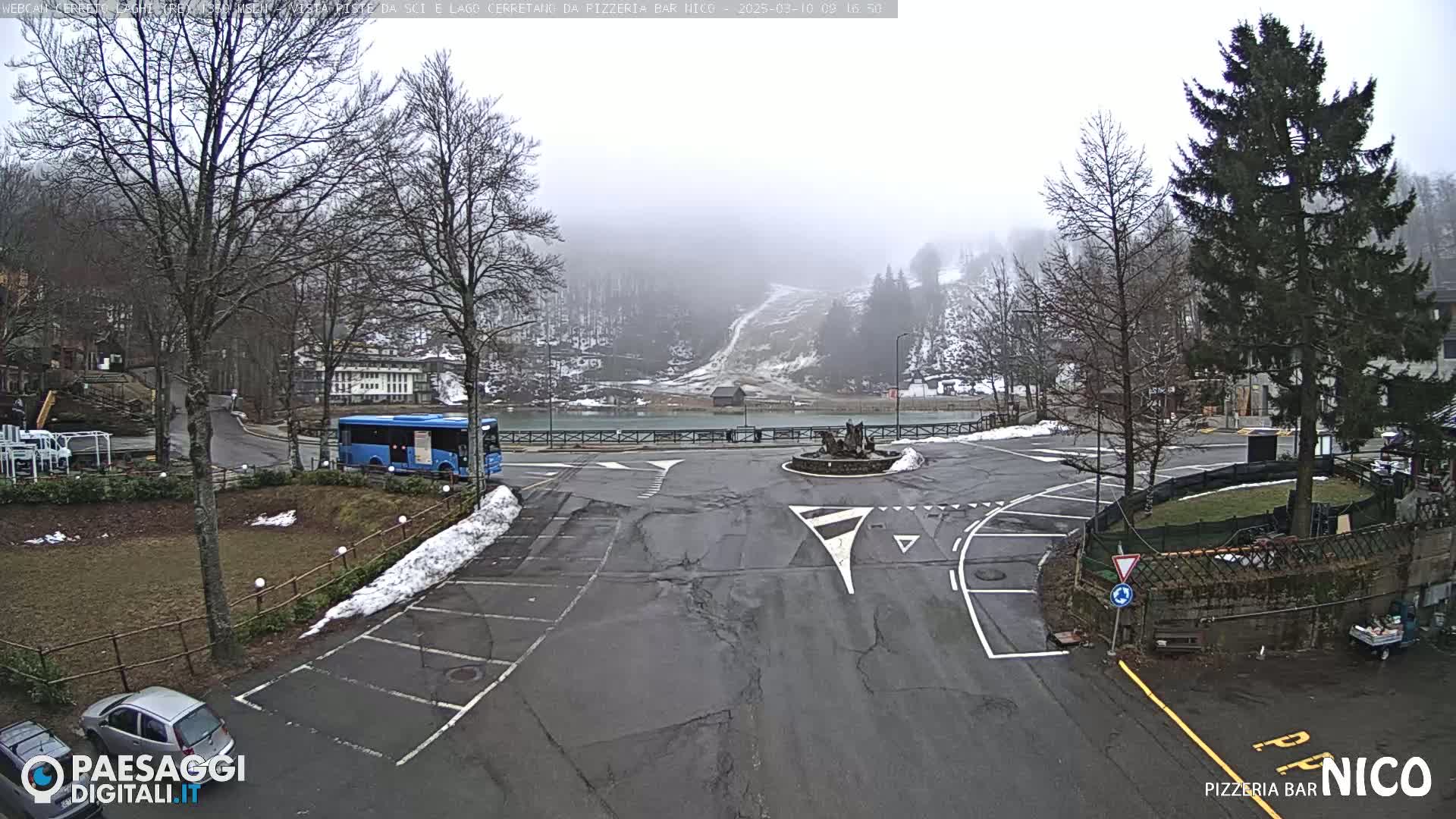 A foggy parking lot with a blue bus, several cars, and trees, near a lake and snow-covered hills.