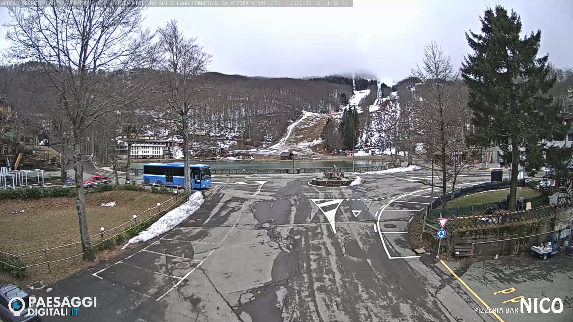 A mostly empty parking lot with a blue bus and some cars, next to a small lake and ski slopes in a wooded area under a cloudy sky.