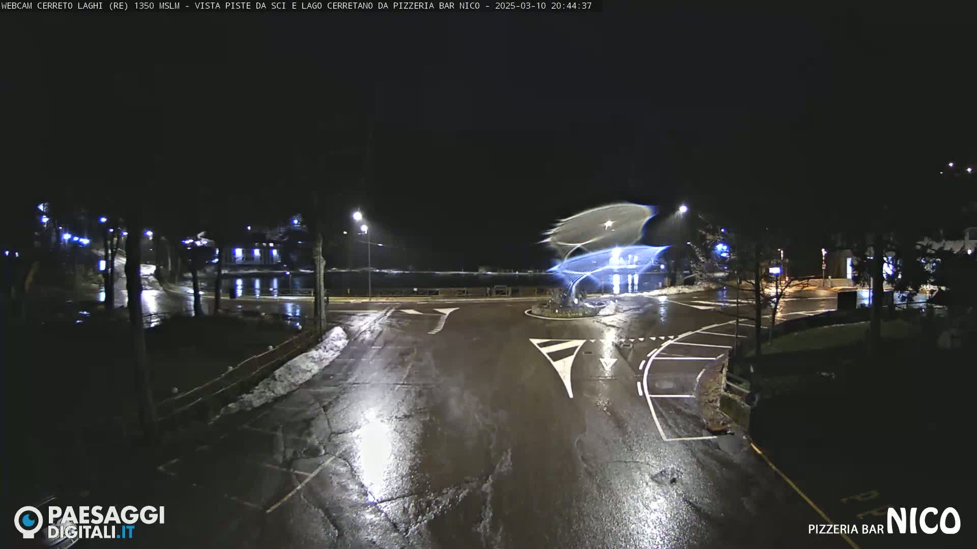A wet, dark night scene shows a roundabout intersection with streetlights, some snow on the ground, and a building in the background.