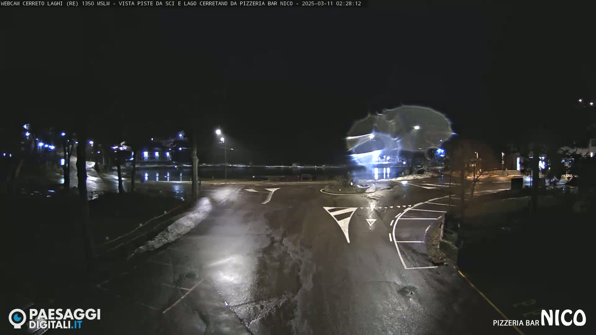 A wet, nighttime street intersection with some snow on the ground and a few streetlights illuminating the scene.