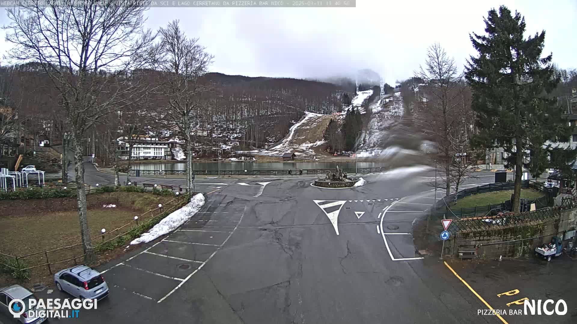 A mostly empty parking lot and roundabout are situated in front of a lake and snow-covered ski slope in overcast weather.