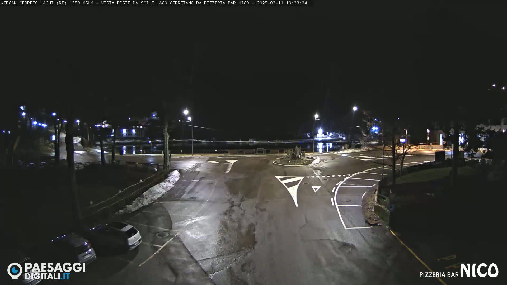 A nighttime view of a wet, dark roadway intersection with some parked cars and streetlights, and a body of water visible in the background.
