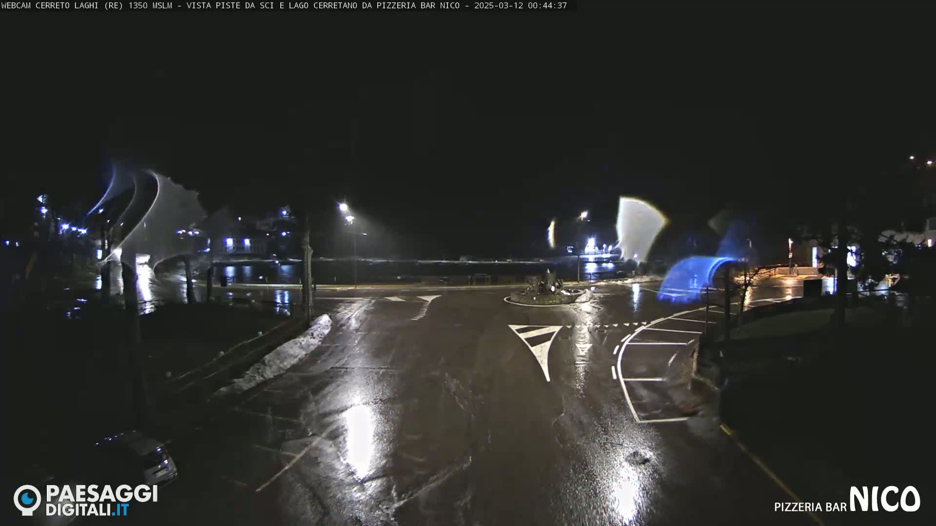 A nighttime, wet road scene shows a roundabout with a parking lot and some buildings in the background, under rainy conditions.