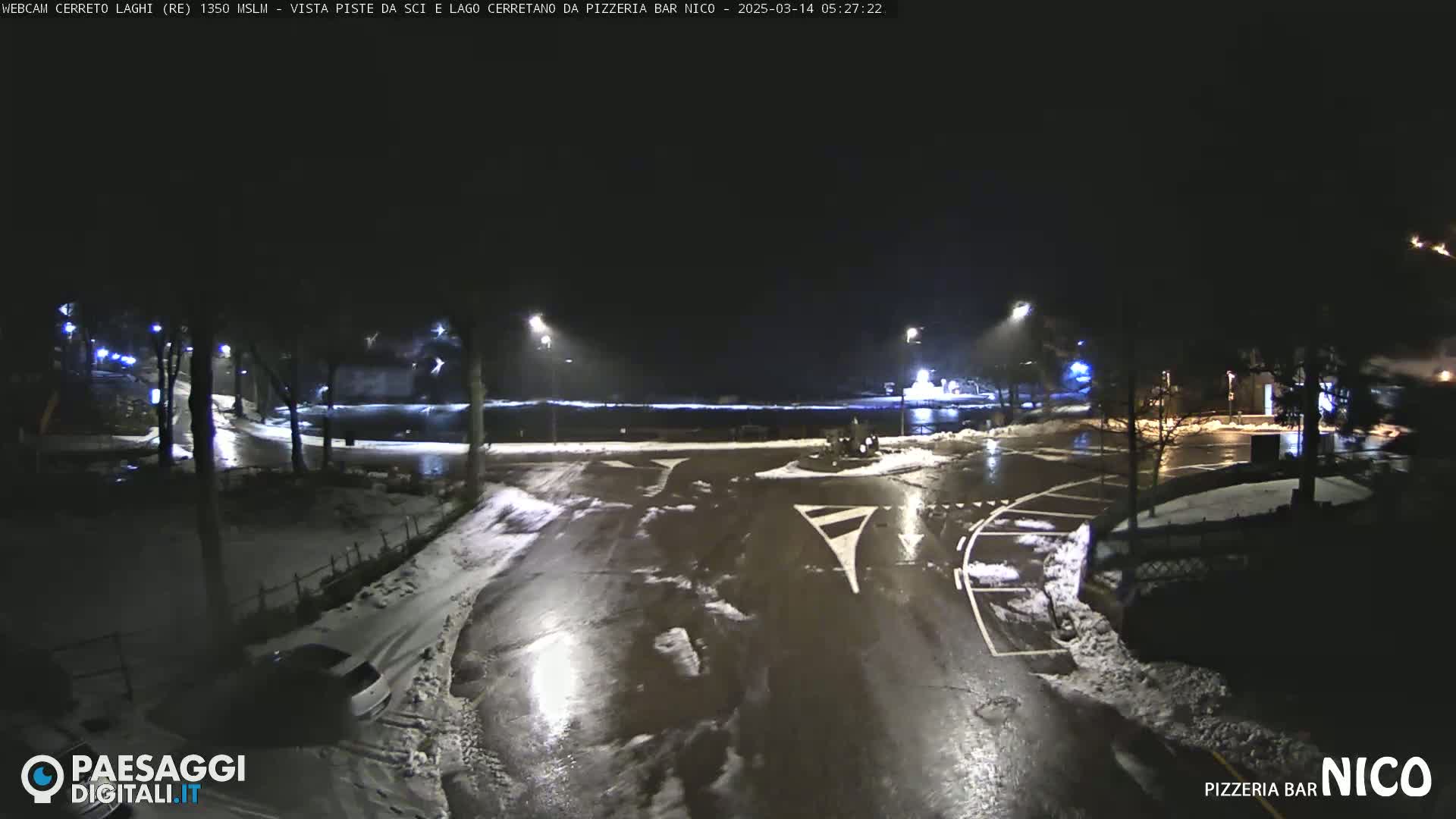 A nighttime view of a snow-dusted parking lot and roadway, illuminated by streetlights, with some wet surfaces.