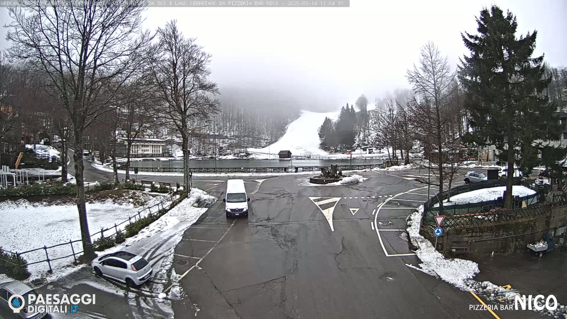 A snowy parking lot with several cars and a van, overlooks a snow-covered ski slope and a small lake, under a foggy sky.