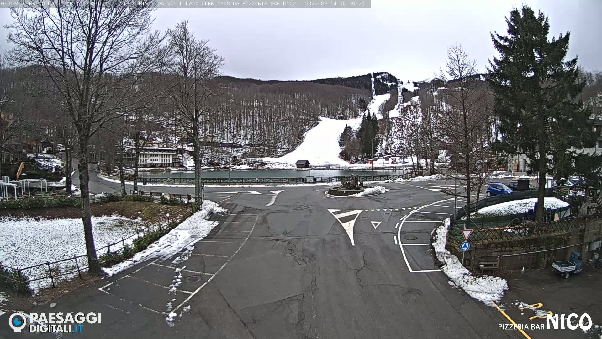 A snow-covered parking lot and roadway are situated near a small lake and a snow-covered ski slope, with several buildings and trees visible in the background under an overcast sky.