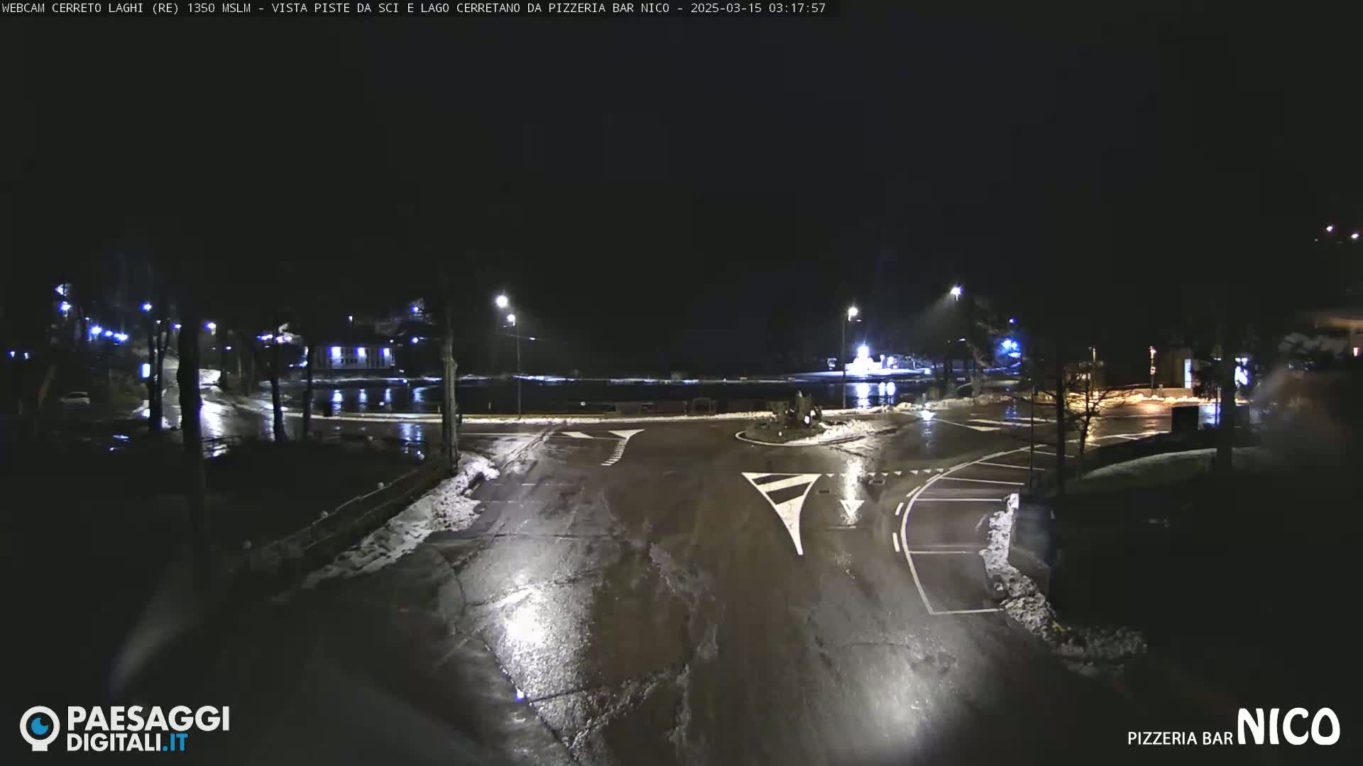 A wet, nighttime view of a roundabout with some snow on the ground and trees illuminated by streetlights.