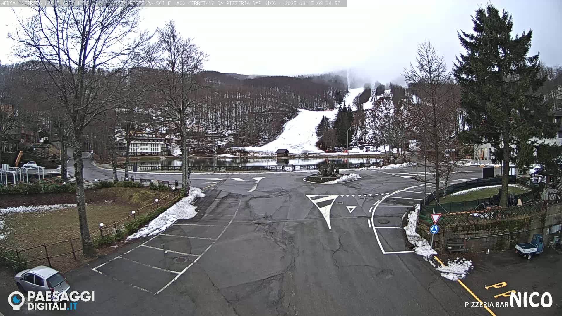A parking lot and roundabout overlook a snow-covered ski slope and a small lake, all under an overcast sky.