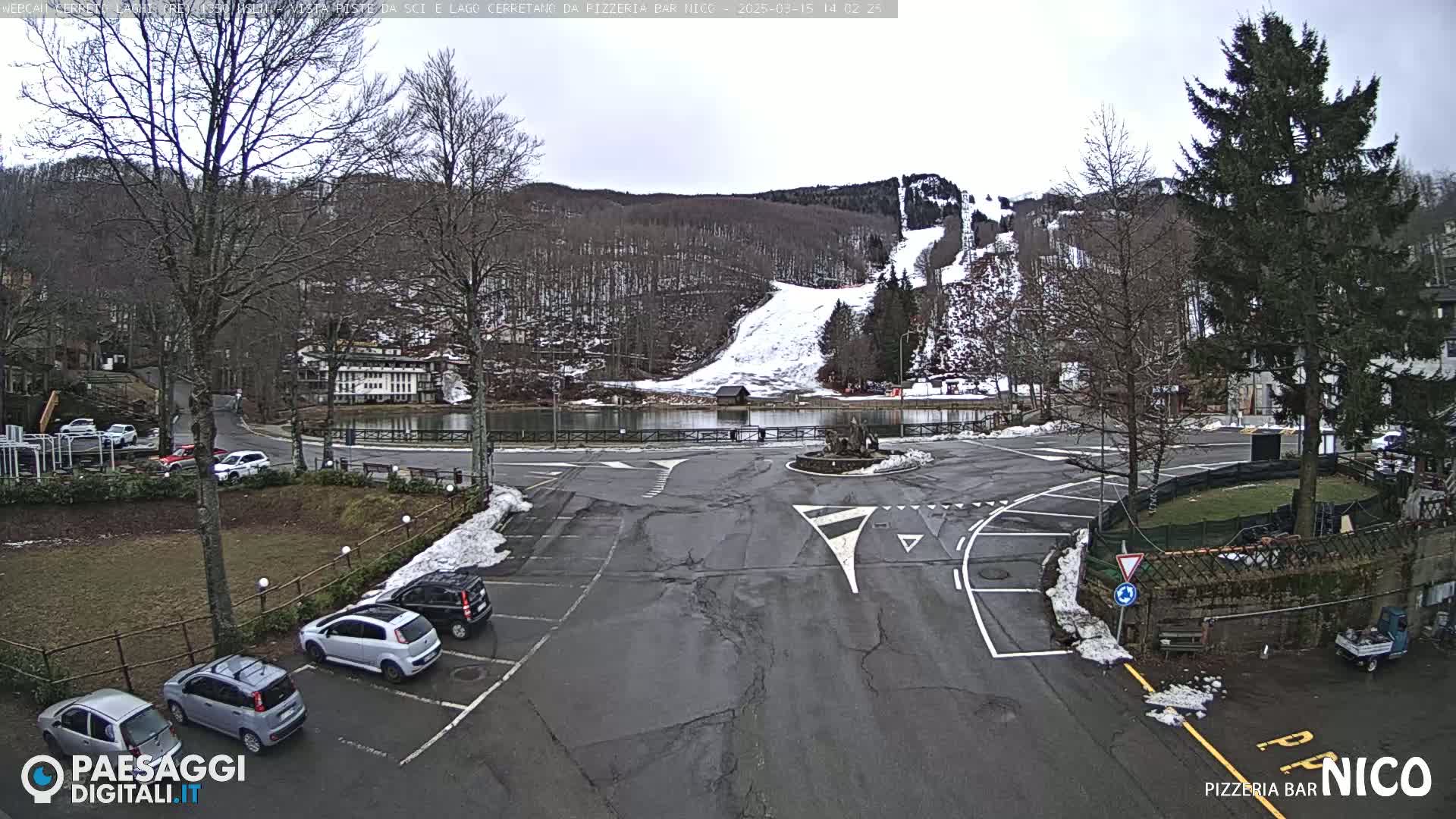 A parking lot with several cars is situated near a small lake, with a ski slope visible in the background on a cloudy day.
