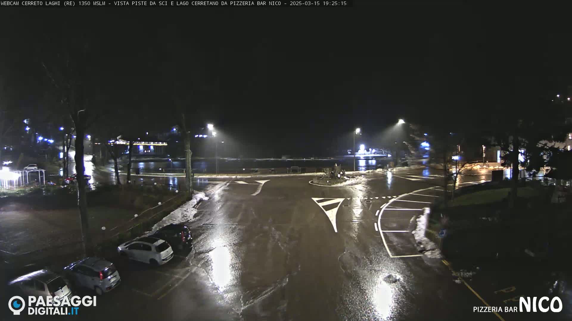 A wet, nighttime scene shows a parking lot and road intersection with several cars parked, illuminated by streetlights and some nearby buildings.
