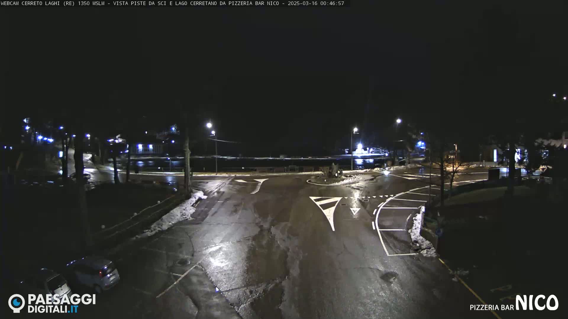 A nighttime view of a wet, snow-dusted parking lot and intersection, illuminated by streetlights.