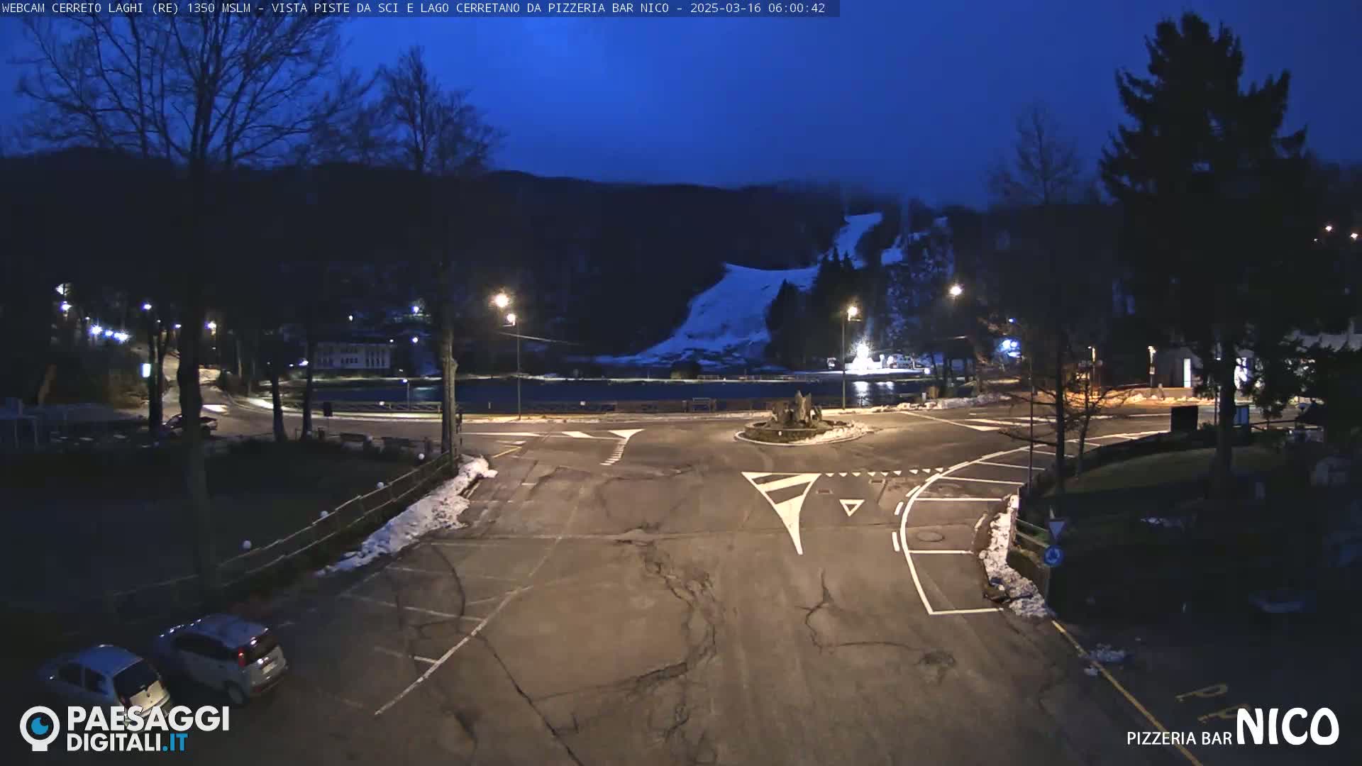 A nighttime view of a parking lot and roadway intersection near a lake, with a snow-covered hill and trees in the background under a dark blue sky.