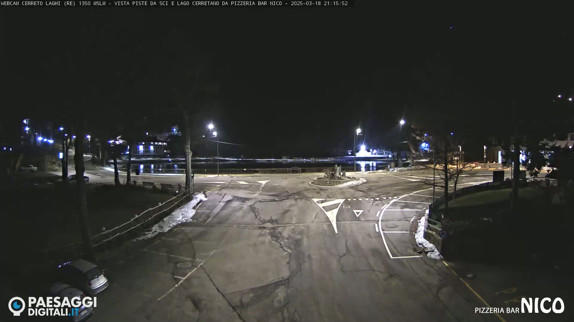 A nighttime view of an empty parking lot and roadway near a body of water, with some snow visible on the ground.