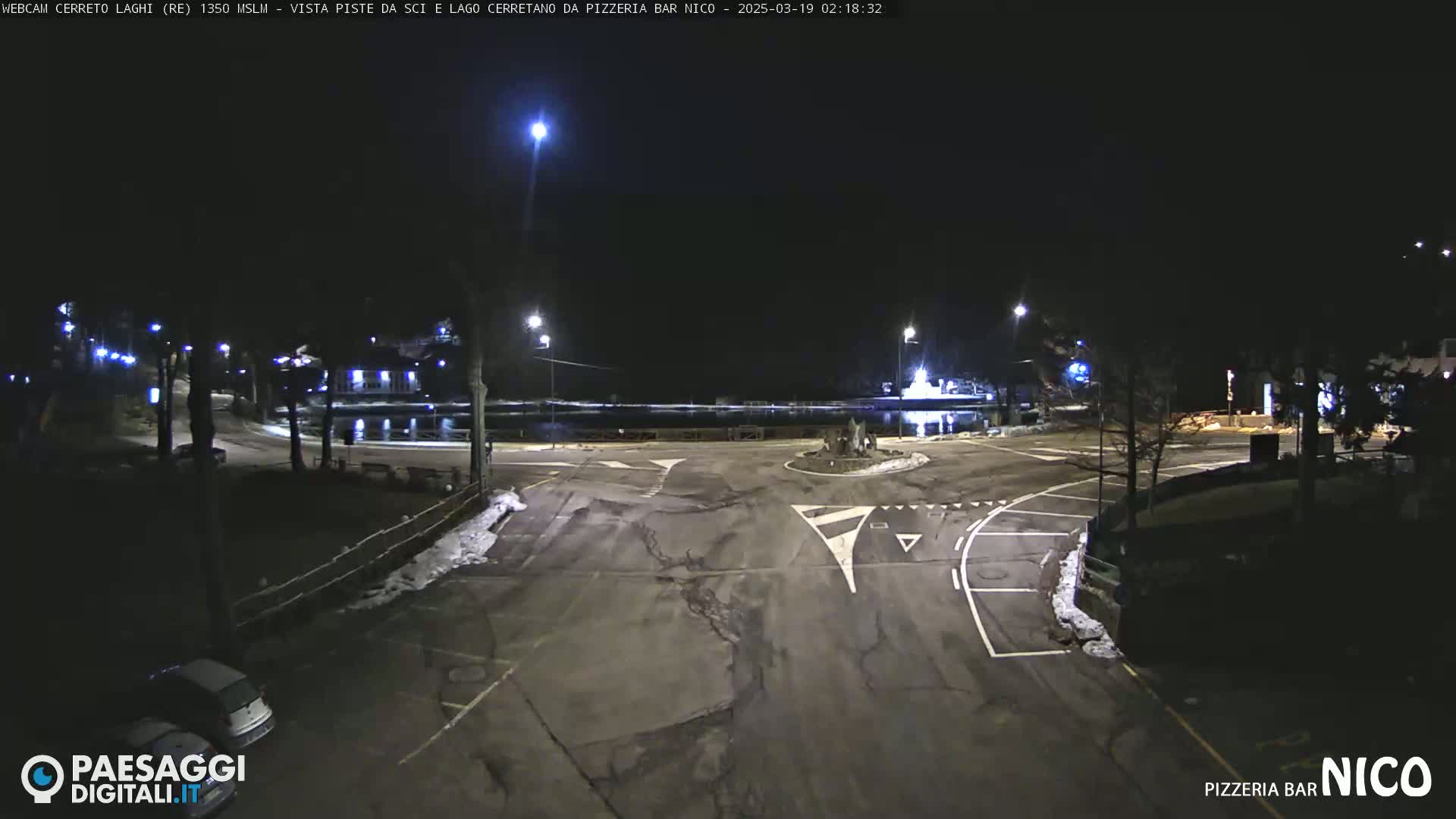 A nighttime view of a mostly empty parking lot and roadway intersection near a body of water, illuminated by streetlights.