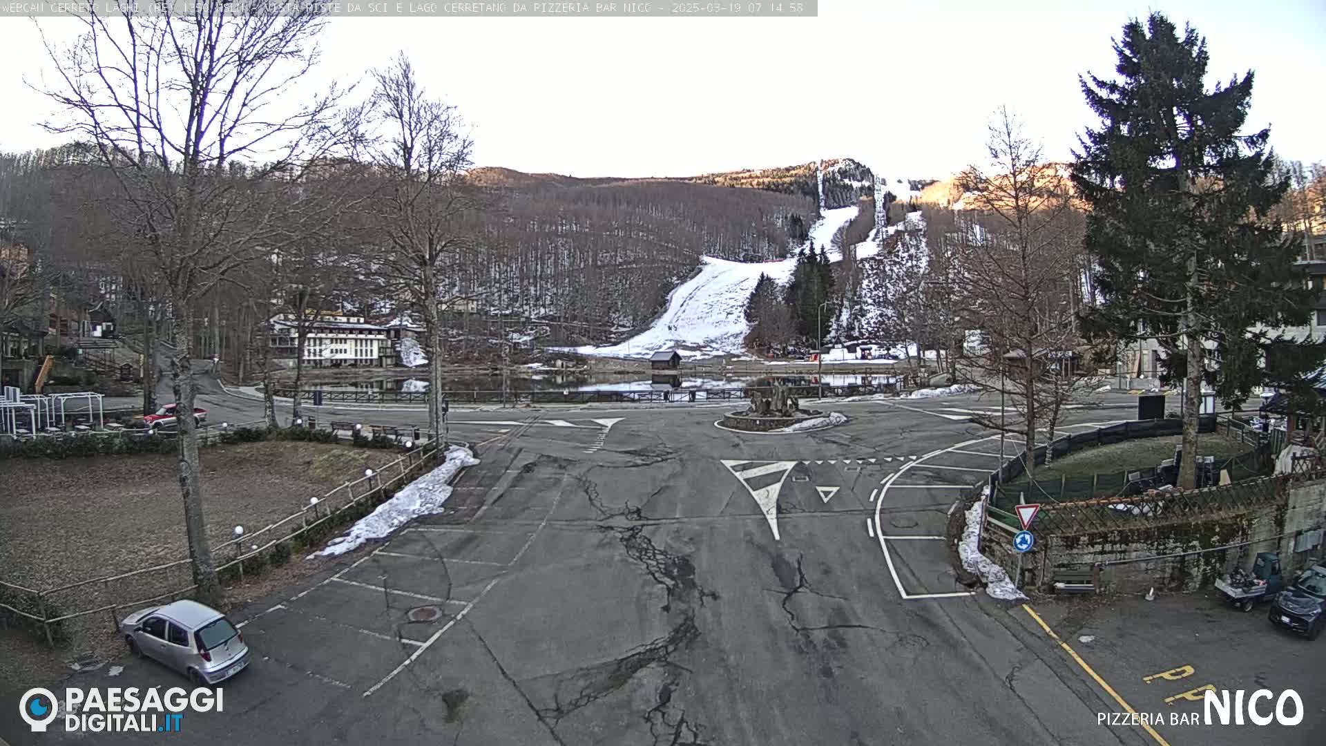 A mostly empty parking lot and road intersection are surrounded by bare trees, a small body of water, and a snow-covered hill in the background under a mostly clear sky.