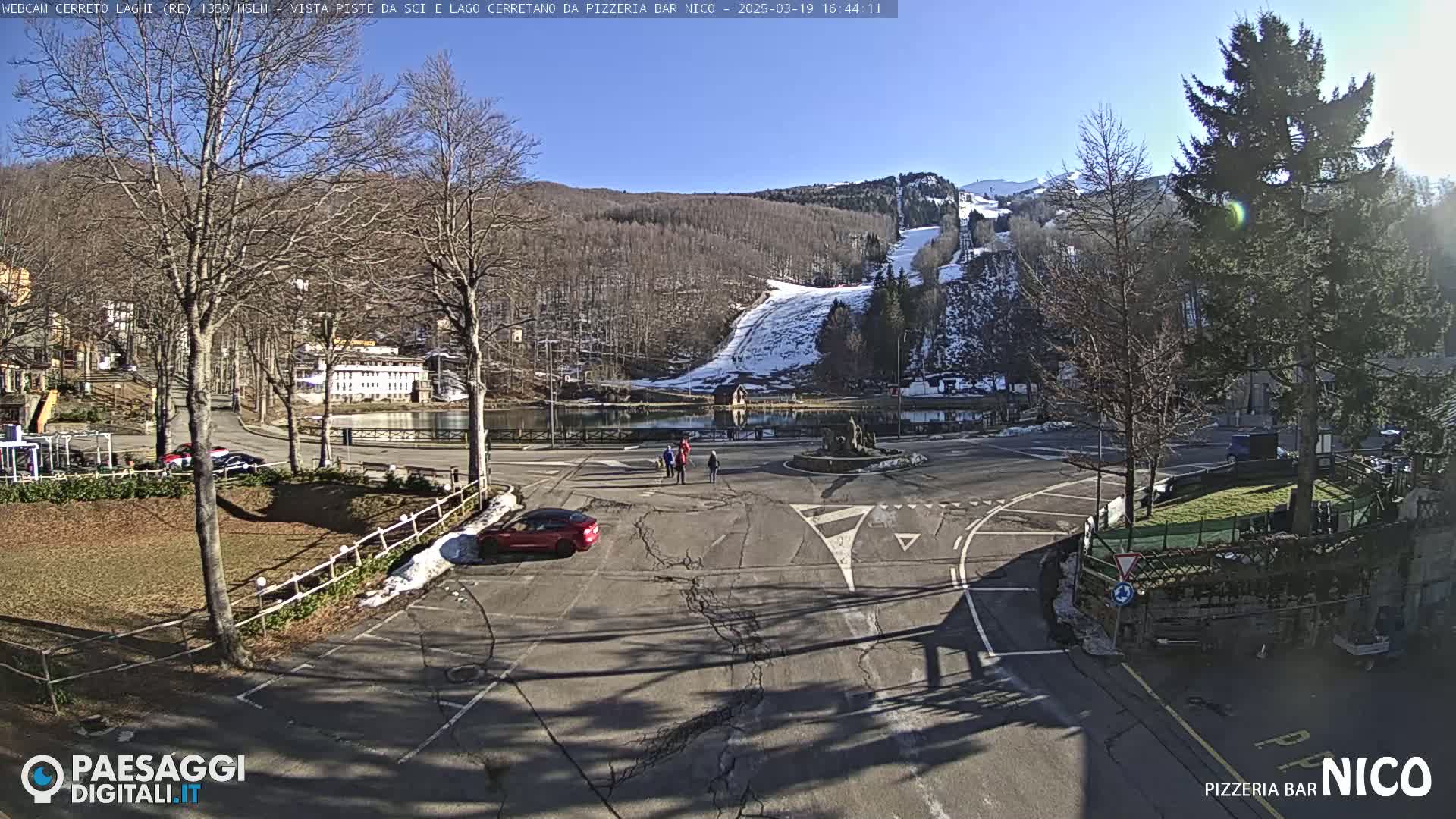 A sunny day reveals a parking lot, a roundabout, a small lake, and snow-covered ski slopes in the background.