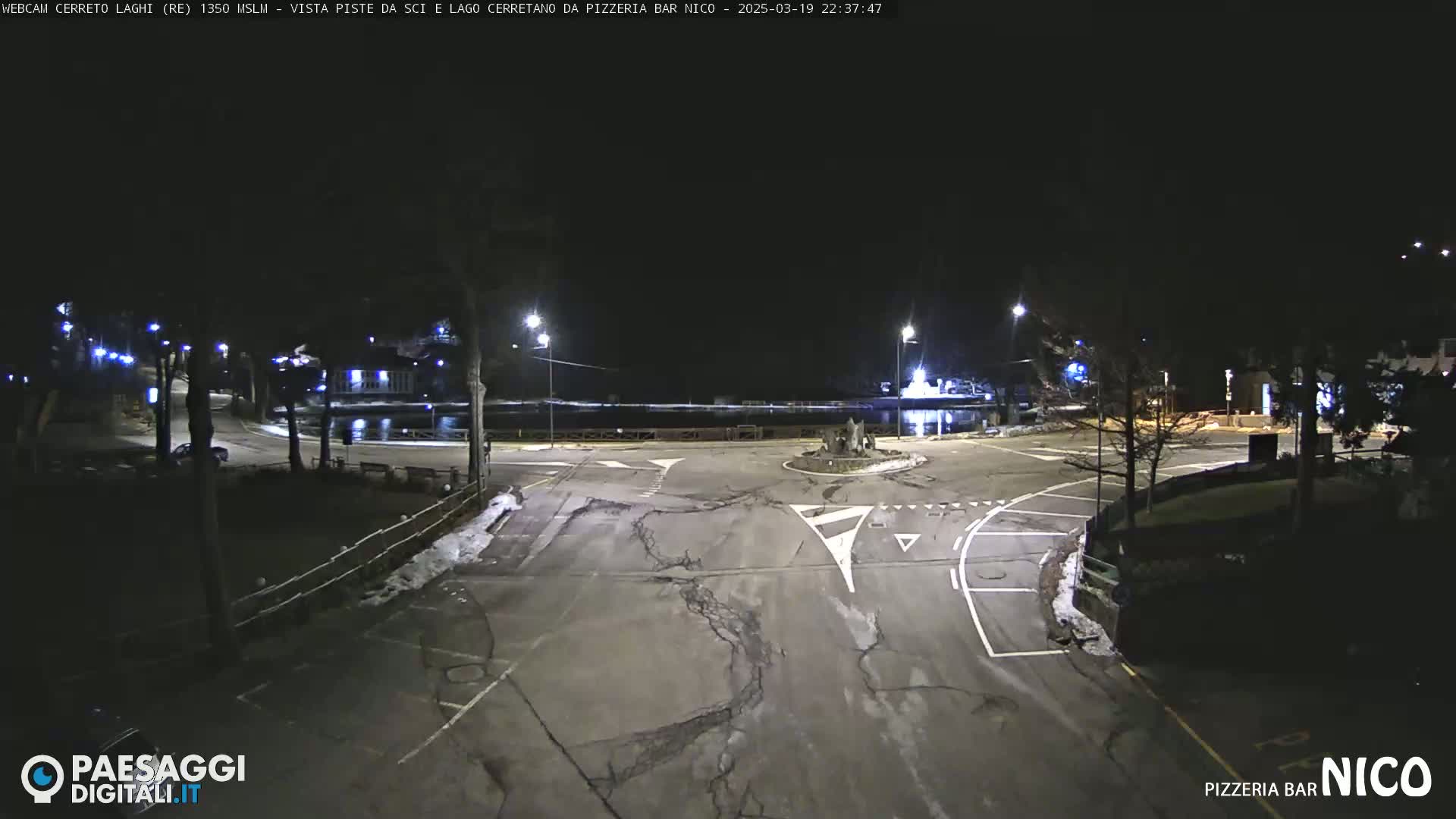 A nighttime, snow-dusted parking lot and roadway intersection are illuminated by streetlights.