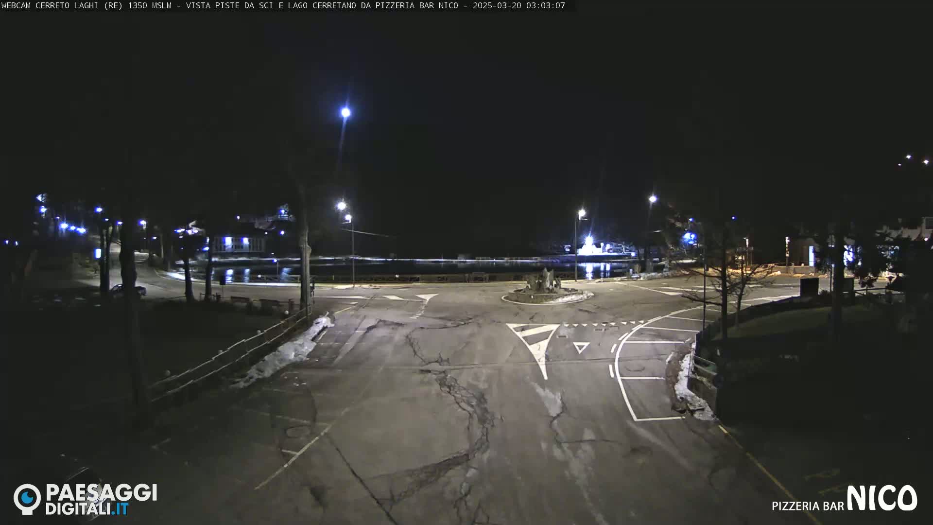 A nighttime view of a paved intersection and parking lot near a body of water, illuminated by streetlights with some snow visible on the ground.