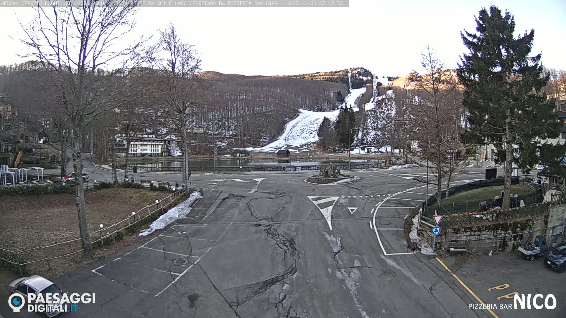 A mostly empty parking lot and roadway surround a small pond, with a snow-covered ski slope and bare deciduous trees visible in the background under a mostly clear sky.